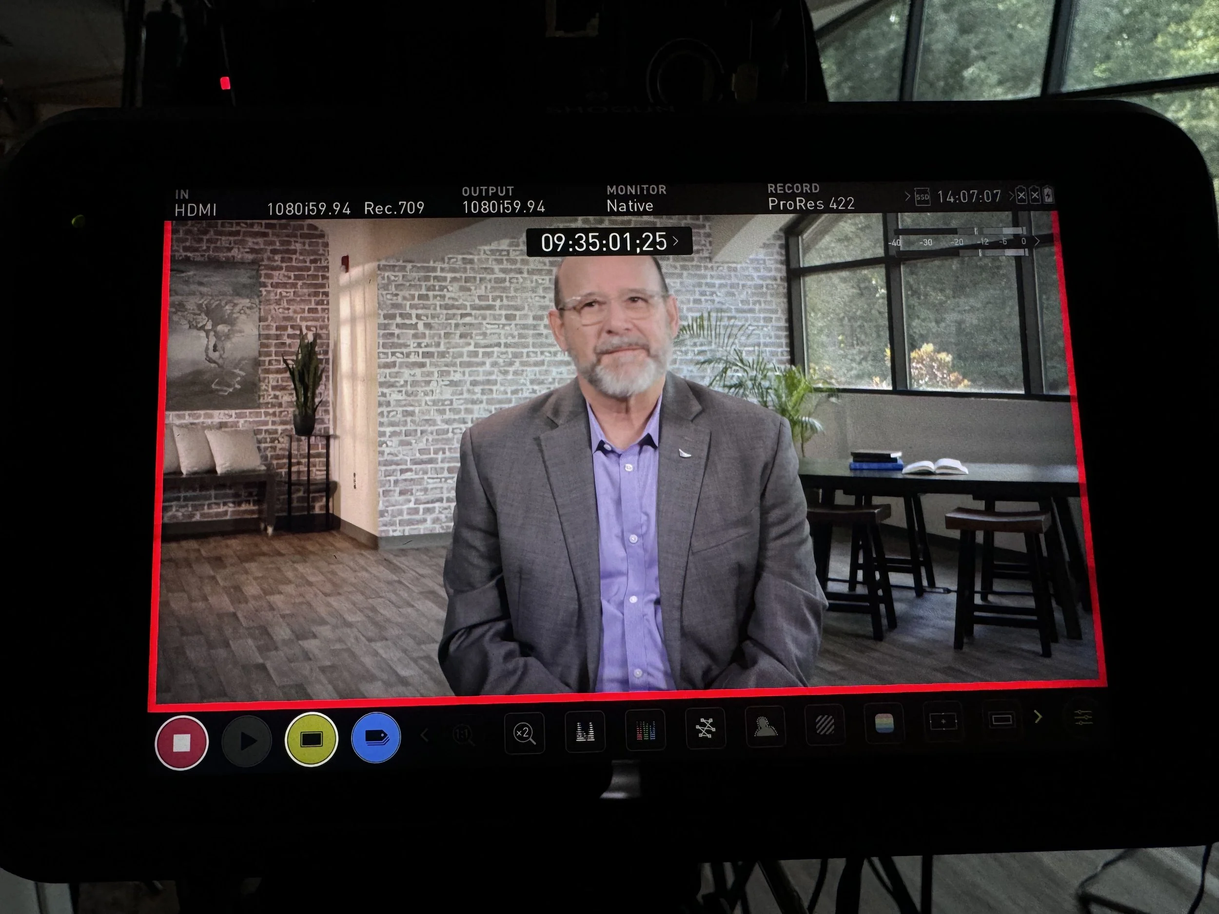 A camera screen shows a man with gray hair and beard wearing glasses and a gray blazer sitting in a room with a brick wall and large windows, with indoor plants and furniture visible in the background.