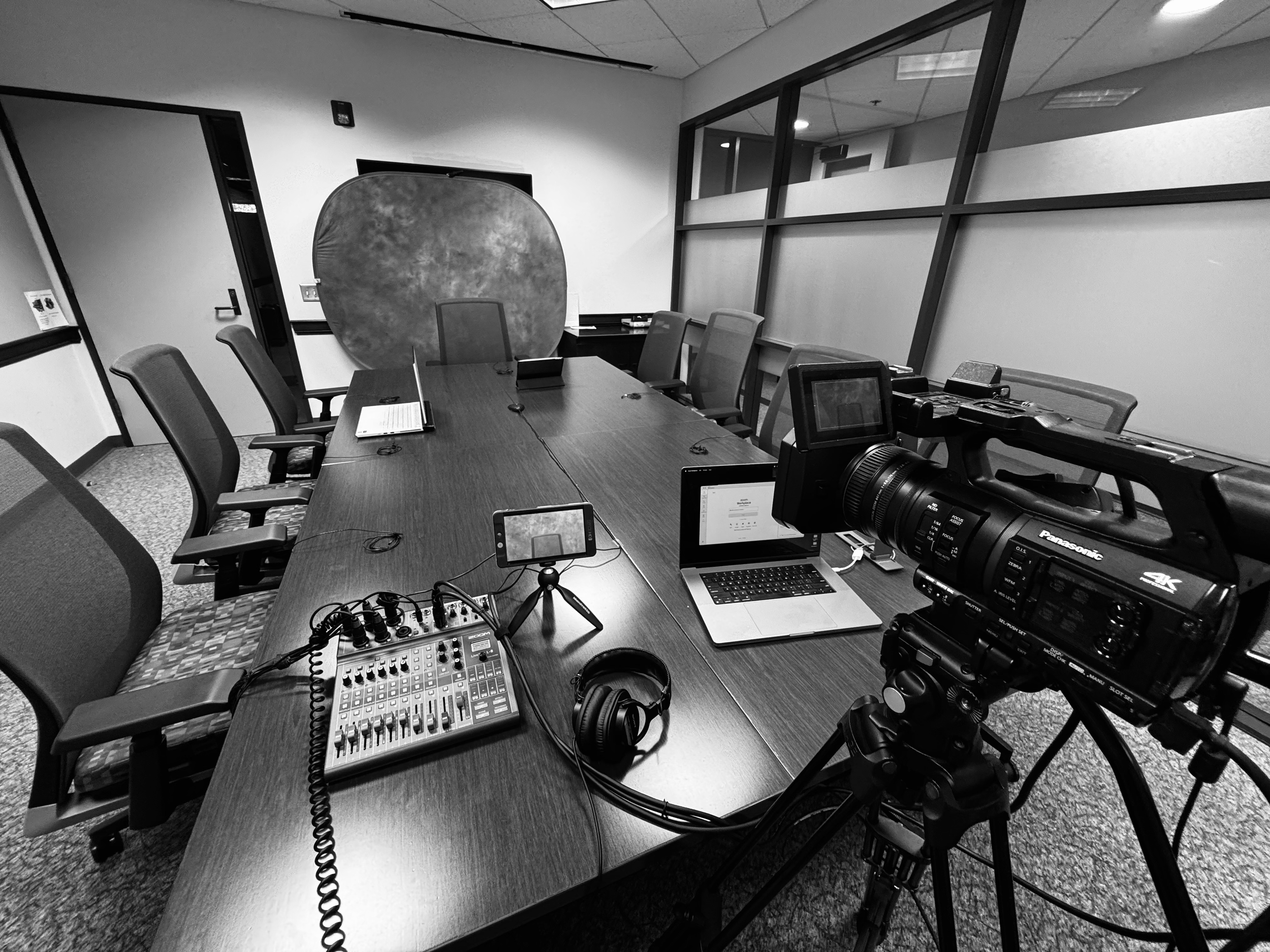 Black and white photo of a conference room with a long table, chairs, a laptop, headphones, a camera, and a video switcher on the table. A large, multi-pane window is on one wall, and a whiteboard and door are visible in the background.