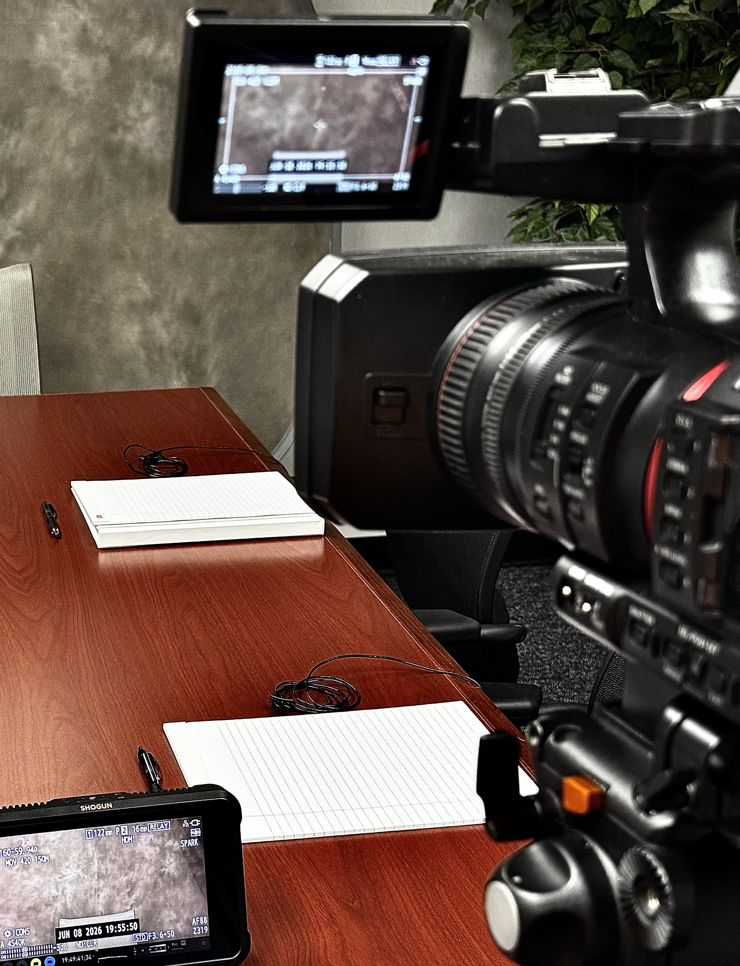 A camera on a tripod capturing a scene of a wooden table with a lined notepad, multiple black earbuds, and pens. There is a monitor and a control device nearby, with a background of a mostly gray wall and green plant leaves.