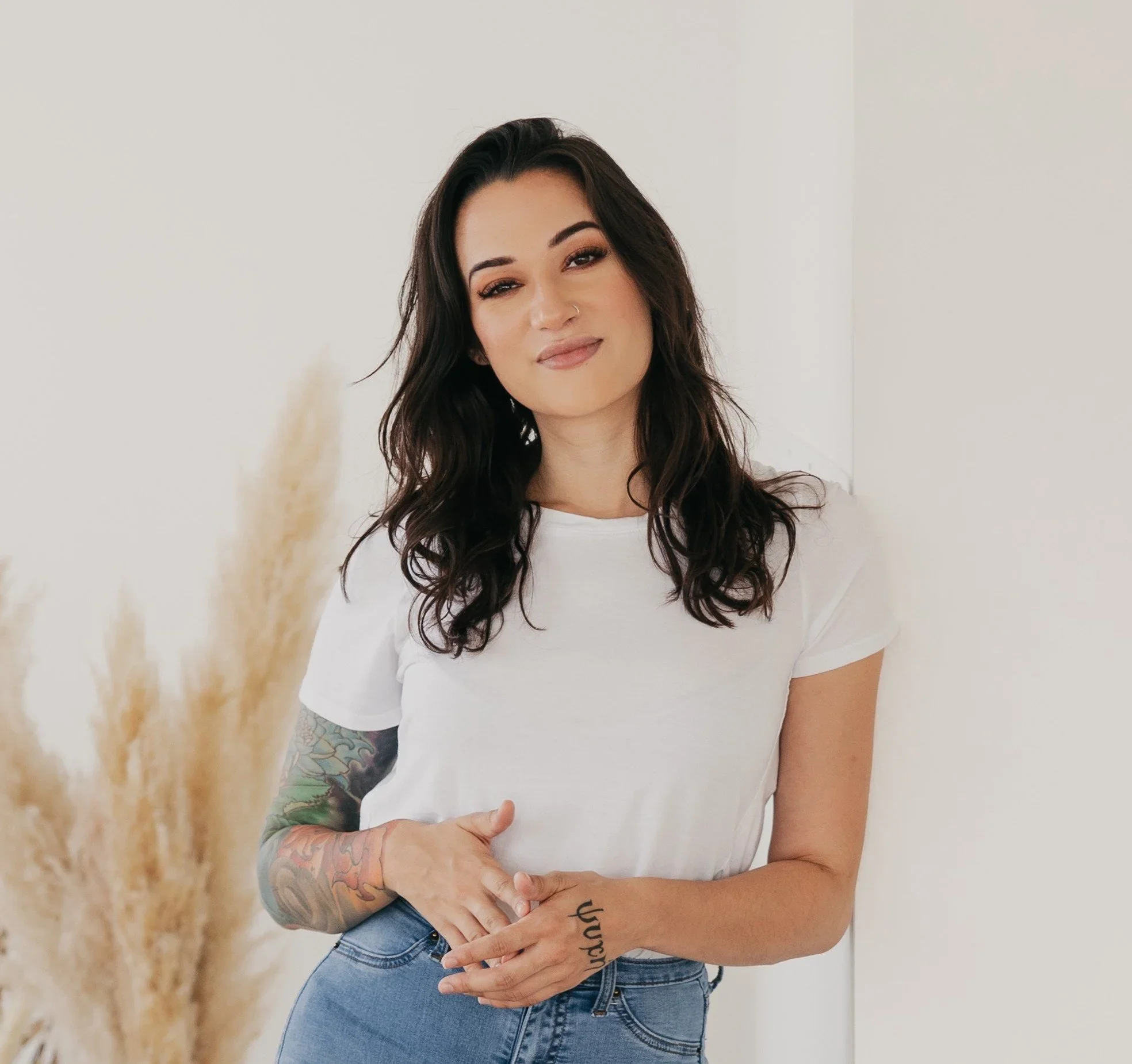 A woman with long dark hair, wearing a white t-shirt and blue jeans, standing against a plain white wall, with a tattooed arm and a tattoo on her hand that says "laugh."