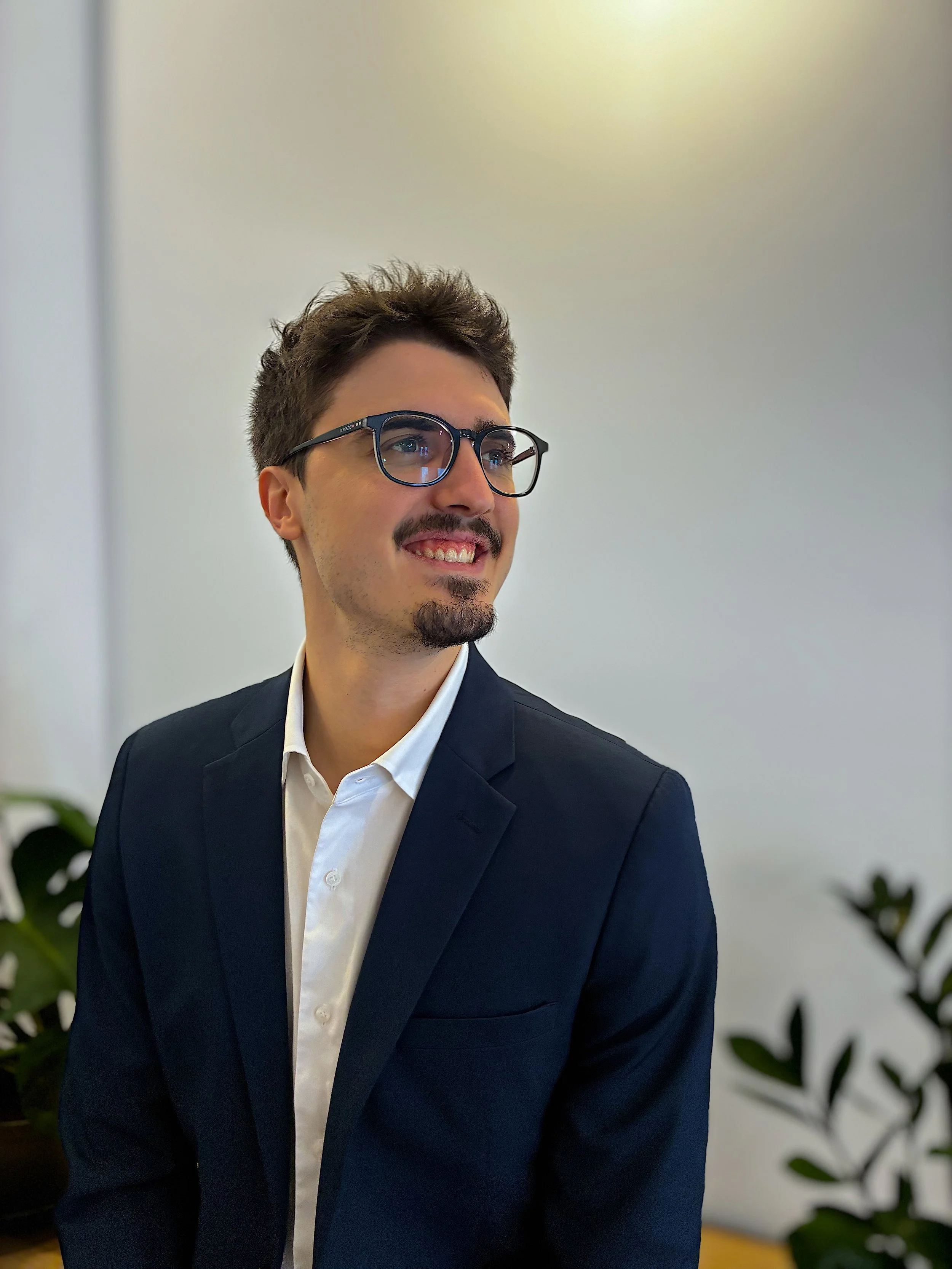 Hombre con barba y gafas, vestido con traje y camisa blanca, sonriendo y mirando hacia la derecha, en un entorno de oficina con plantas verdes.