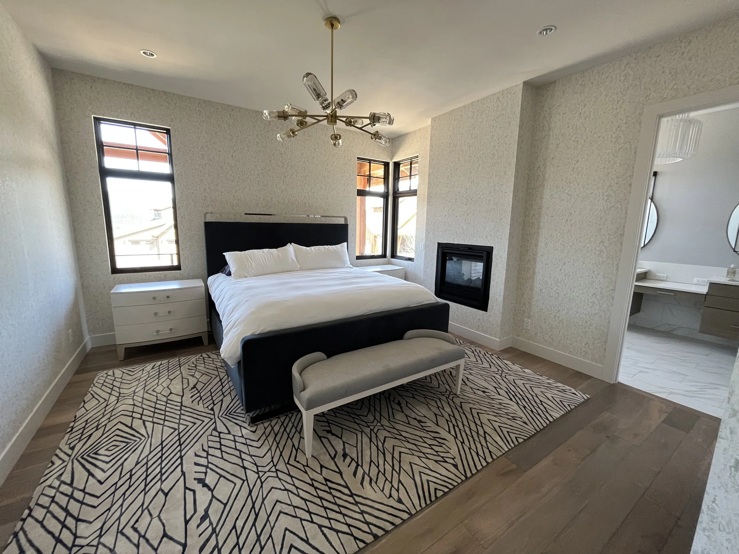 Modern bedroom with a black bed, white bedding, nightstand, large windows, and a black fireplace. Decorated with a patterned rug and a sputnik chandelier.