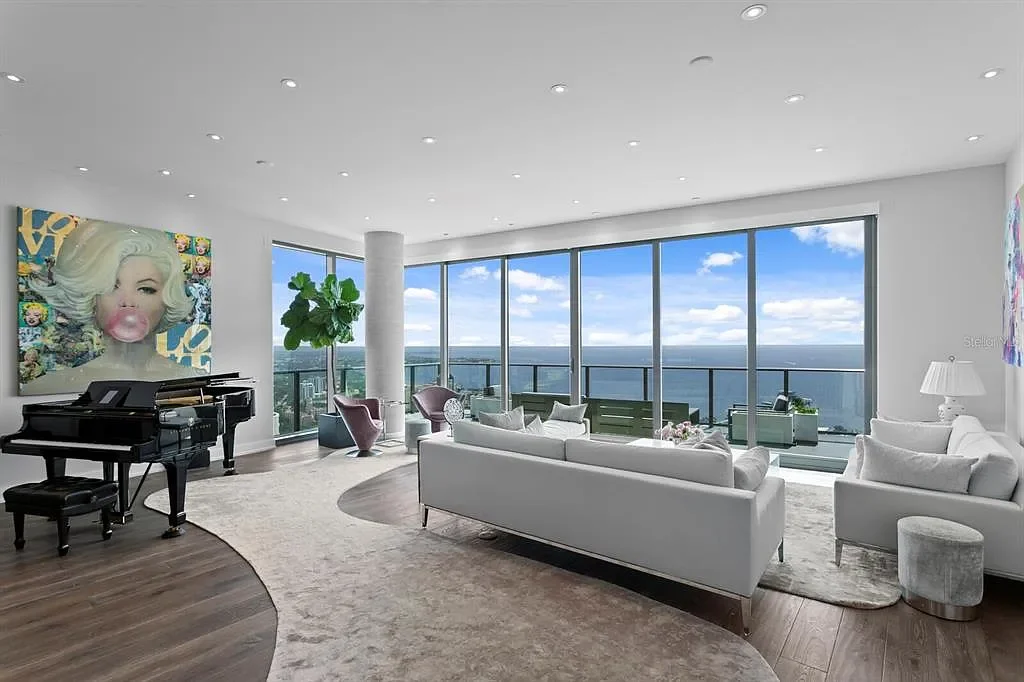 Modern living room with large floor-to-ceiling windows offering a city view, white sofas, a black grand piano, colorful artwork, potted plants, and a mix of contemporary and eclectic decor.