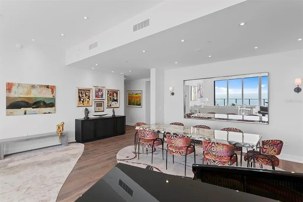 Modern living and dining room with white walls, hardwood floors, colorful artwork, a dining table with red patterned chairs, and a large window viewing an ocean and blue sky.