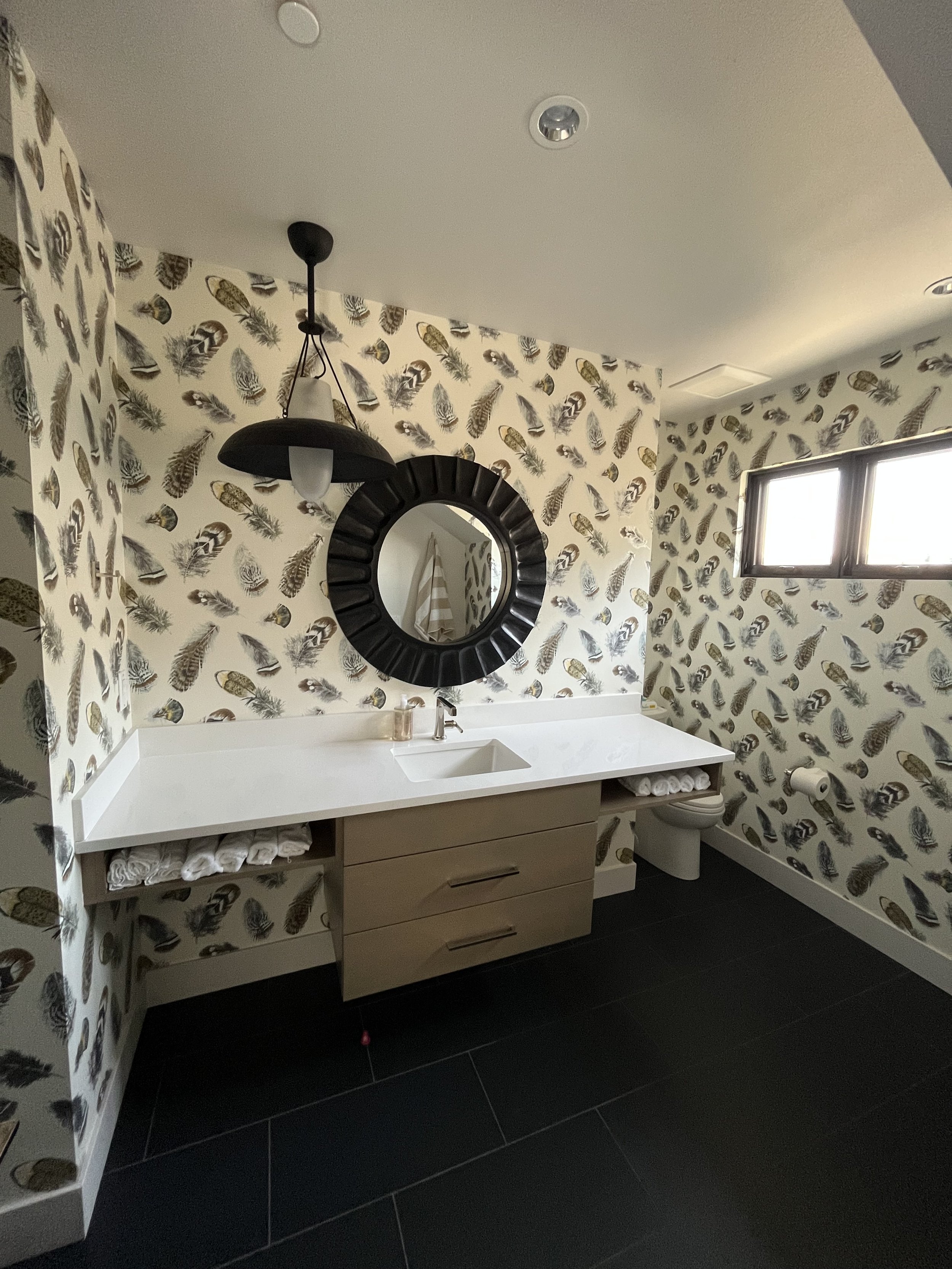 Modern bathroom with black feather print wallpaper, white vanity, round mirror, black pendant light, window, and dark flooring.