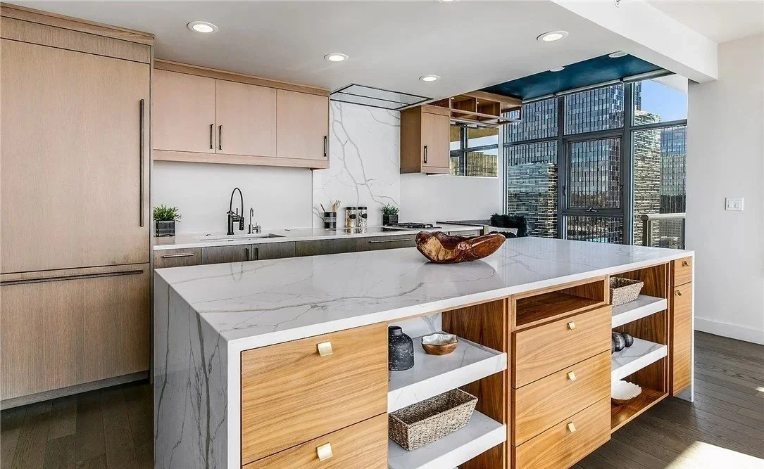 Modern kitchen with light wood cabinets, white marble countertop and a large island. High-rise buildings visible through floor-to-ceiling windows.
