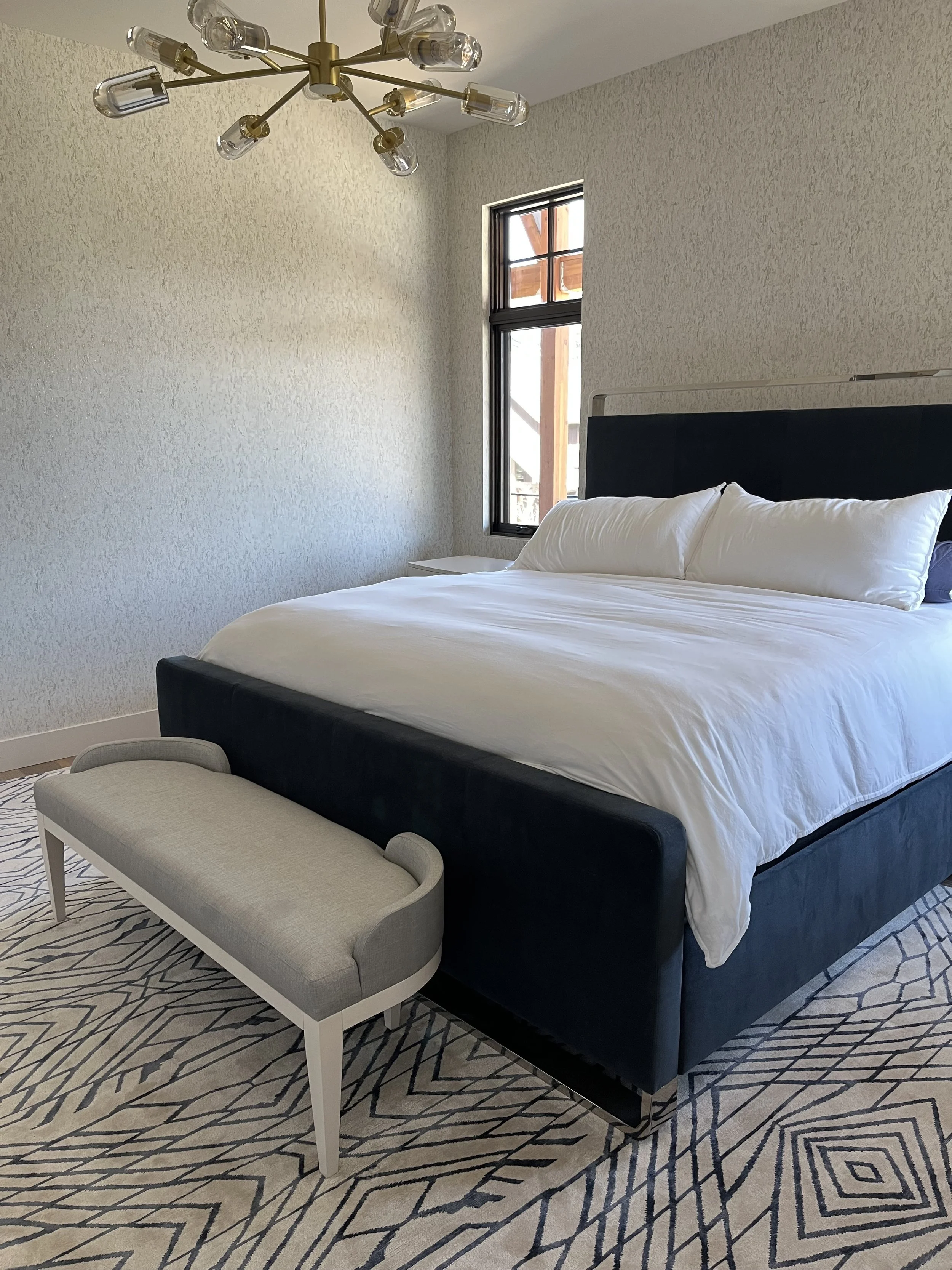 A bedroom with a bed, a bench at the foot of the bed, a window, a textured wall, and a modern chandelier with multiple glass bulbs.