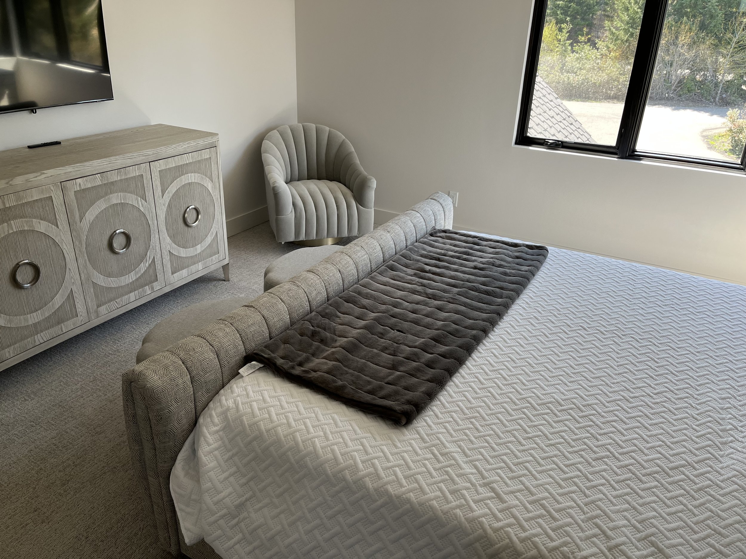 A bedroom with a light-colored upholstered bed, a gray blanket, a white quilted bedspread, a beige patterned armchair, a wooden cabinet with circular details, a wall-mounted TV, and a large window showing greenery outside.