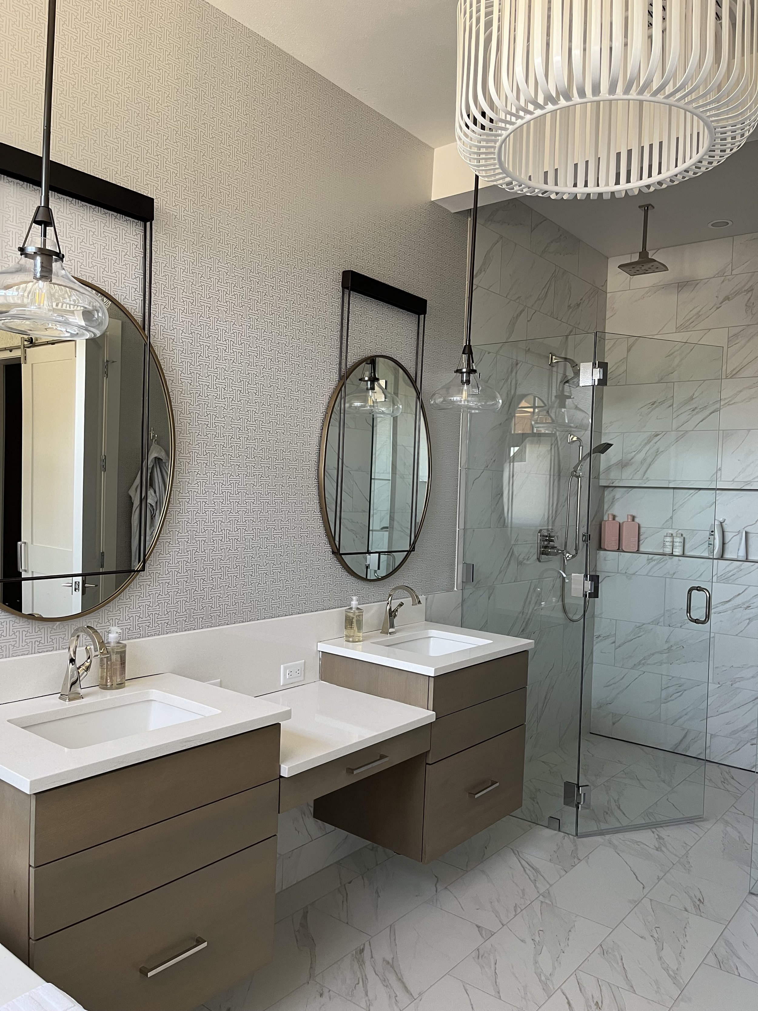 Modern bathroom with dual sinks, oval mirrors, and a glass-enclosed shower with marble tiles and built-in shelves holding pink bottles.