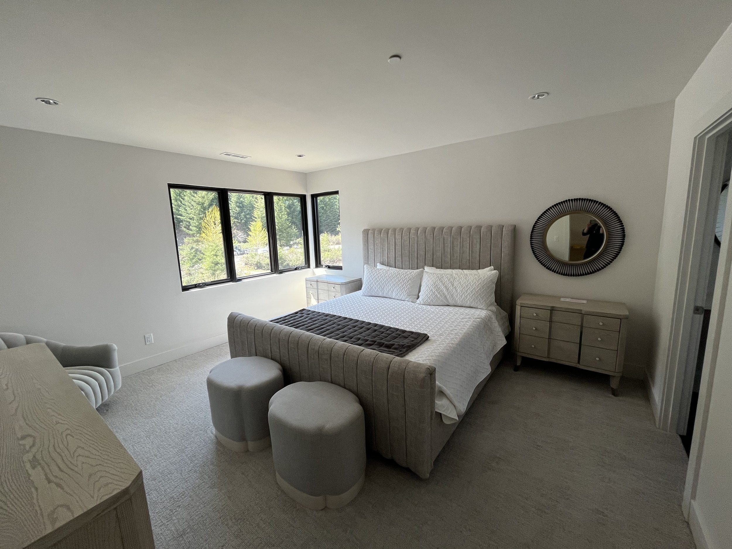 A bedroom with a bed, a round mirror on the wall, a window with trees outside, a nightstand, and two small upholstered stools at the foot of the bed.