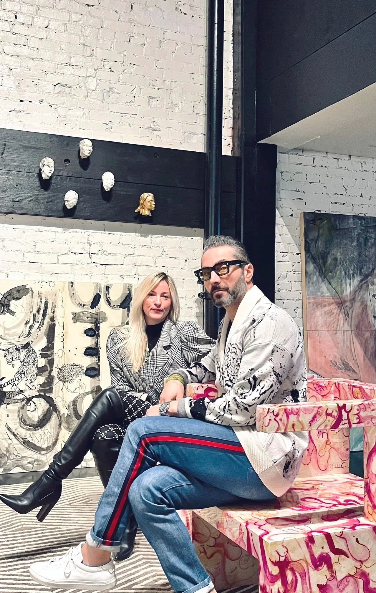 A man and woman sitting on a pink and white patterned bench in an art gallery. The woman has long blonde hair, wearing a black and white checkered outfit and black boots. The man has short gray hair, beard, glasses, wearing a white jacket with black patterns and blue jeans with red stripes. There are artwork and masks on the wall behind them.