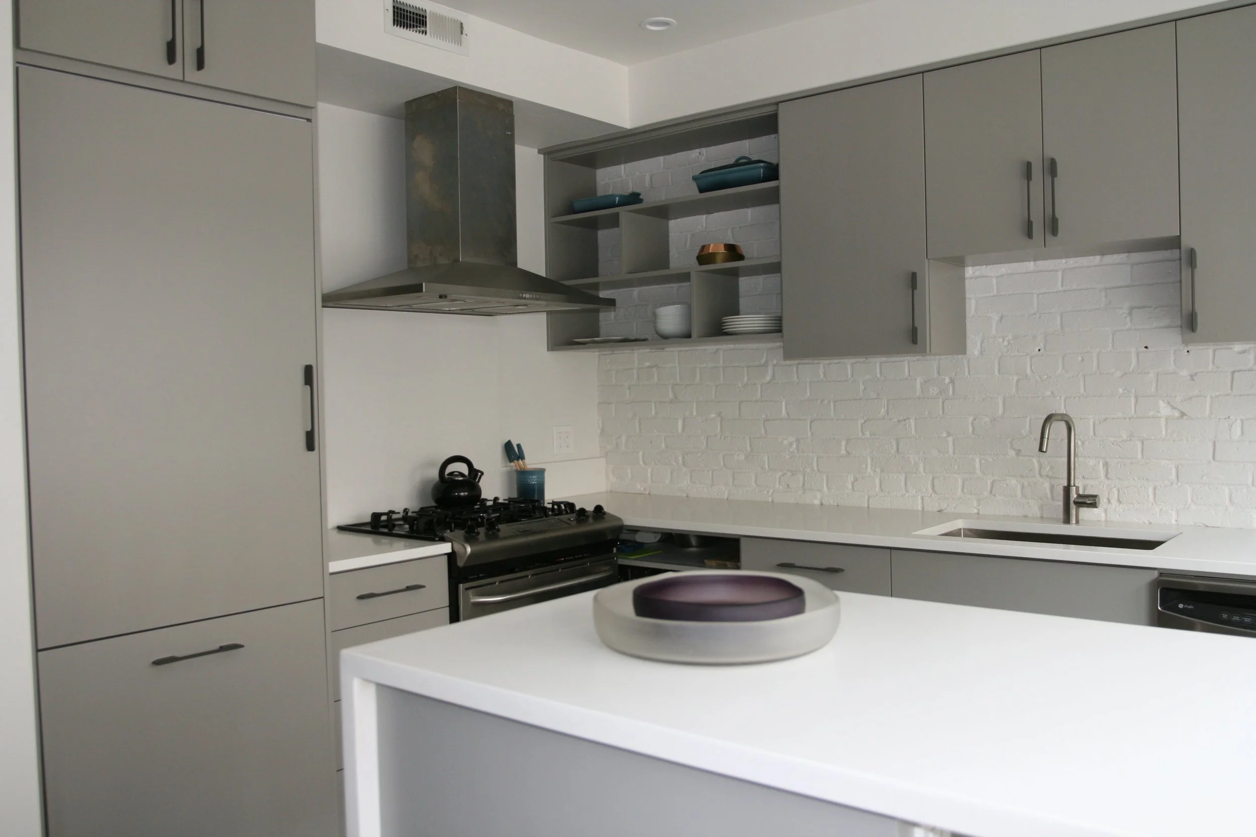 Modern kitchen with gray cabinets, white countertop, brick wall, stainless steel range hood, stove, sink, and dishes.