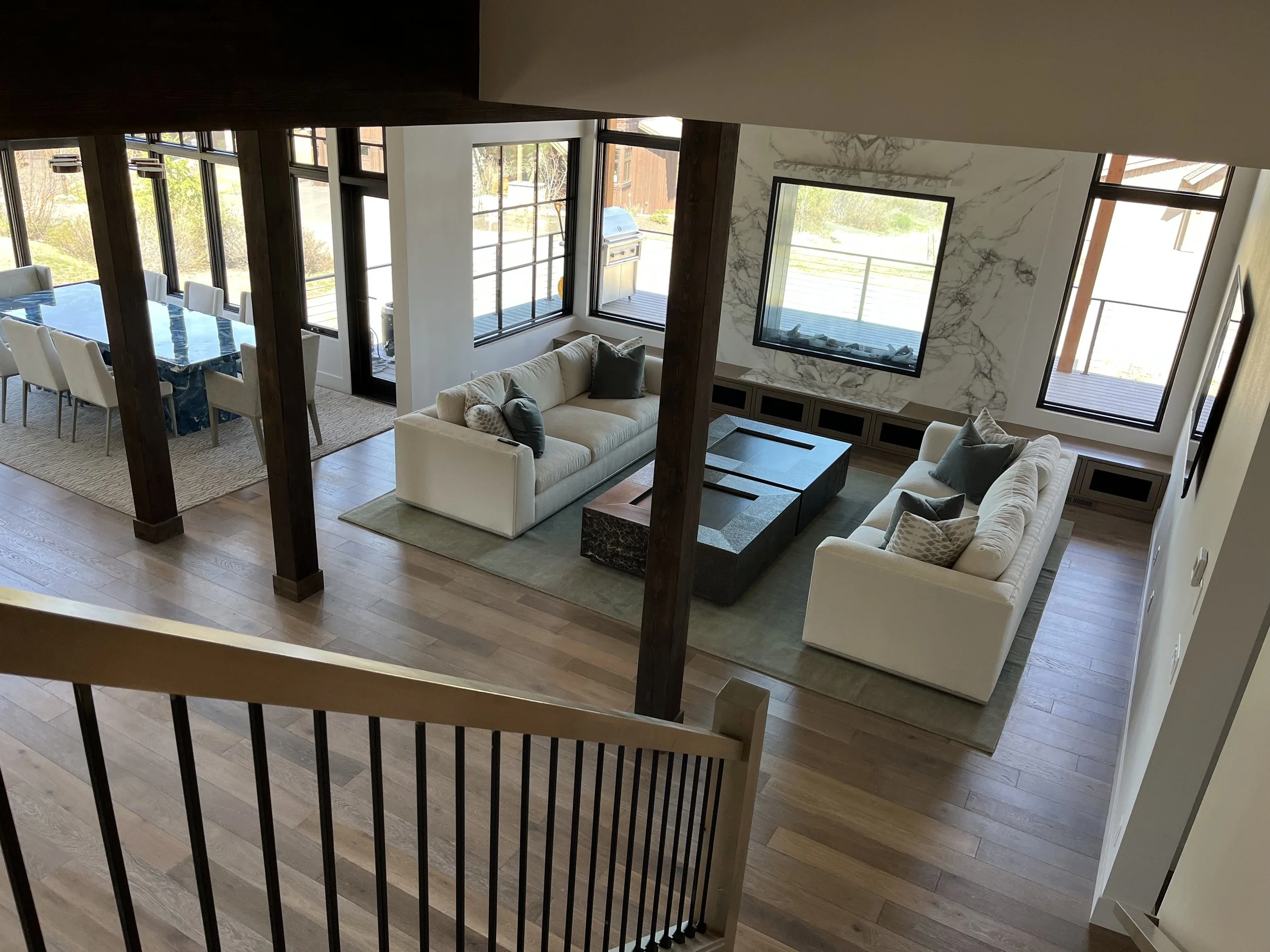 Indoor living space with a beige sofa, multiple chairs, large windows, a marble accent wall, and a view of a deck outside.