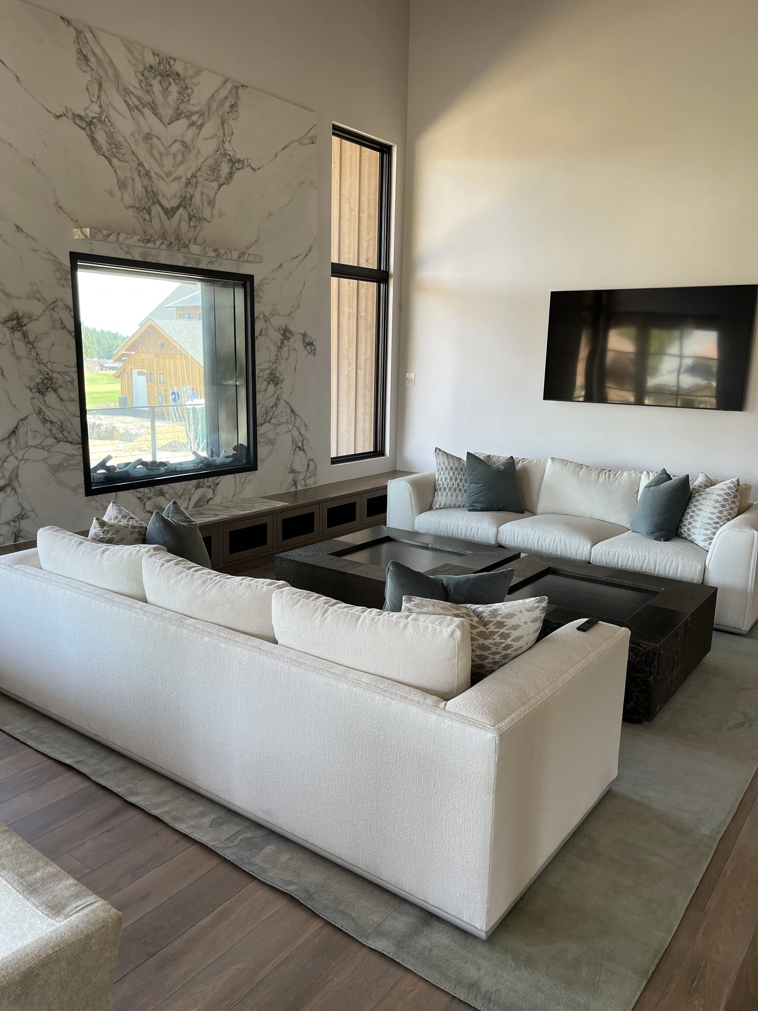 Living room with a large white sectional sofa with various gray and patterned pillows, a black coffee table, a marble accent wall with two windows, and a wall-mounted flat-screen TV.