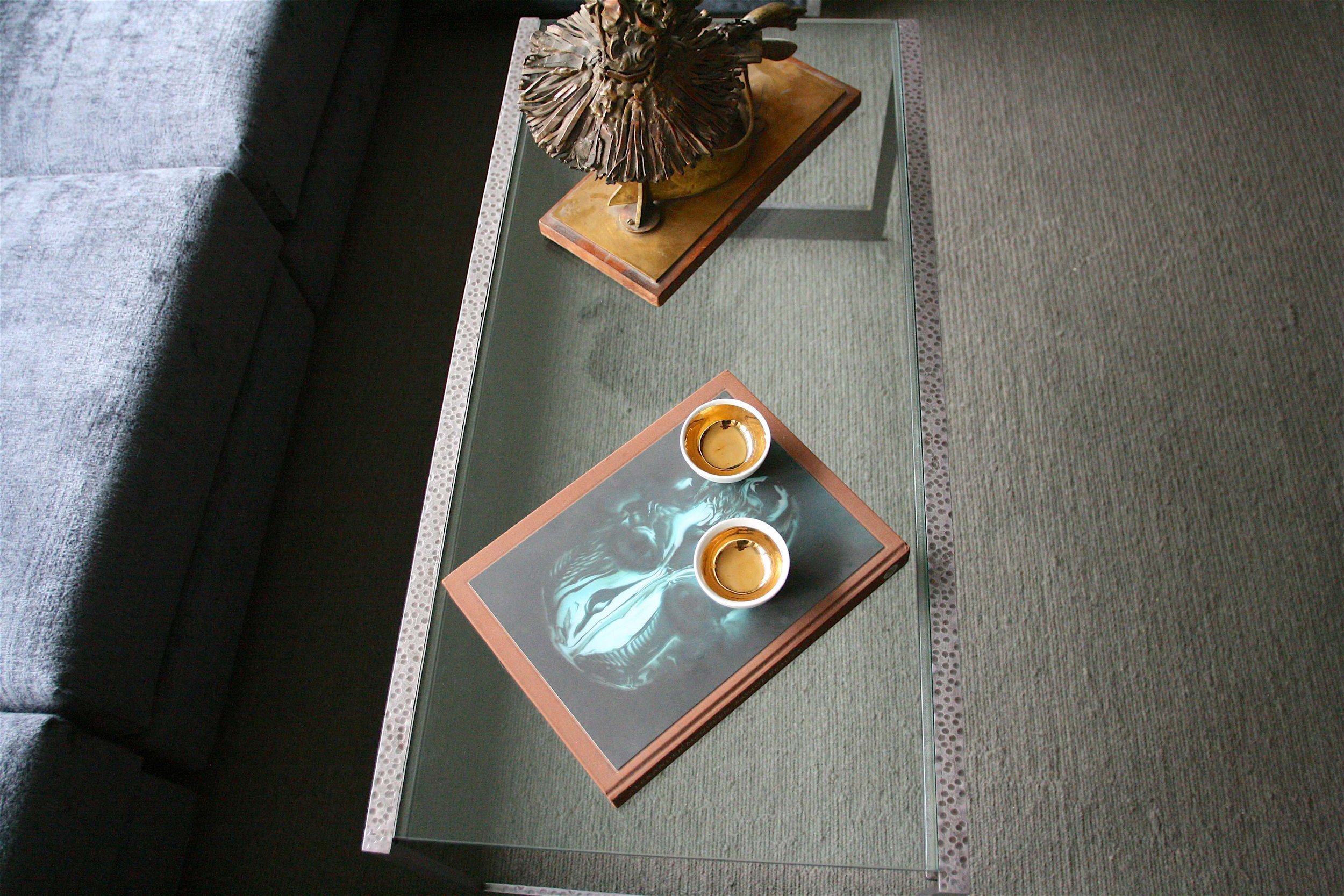 A glass-top coffee table with a wooden base featuring a swirling design, placed in front of a gray upholstered sofa. On the table are two gold-colored bowls and a decorative brown sculpture resembling a twisted tree or coral. The table is on a greeni