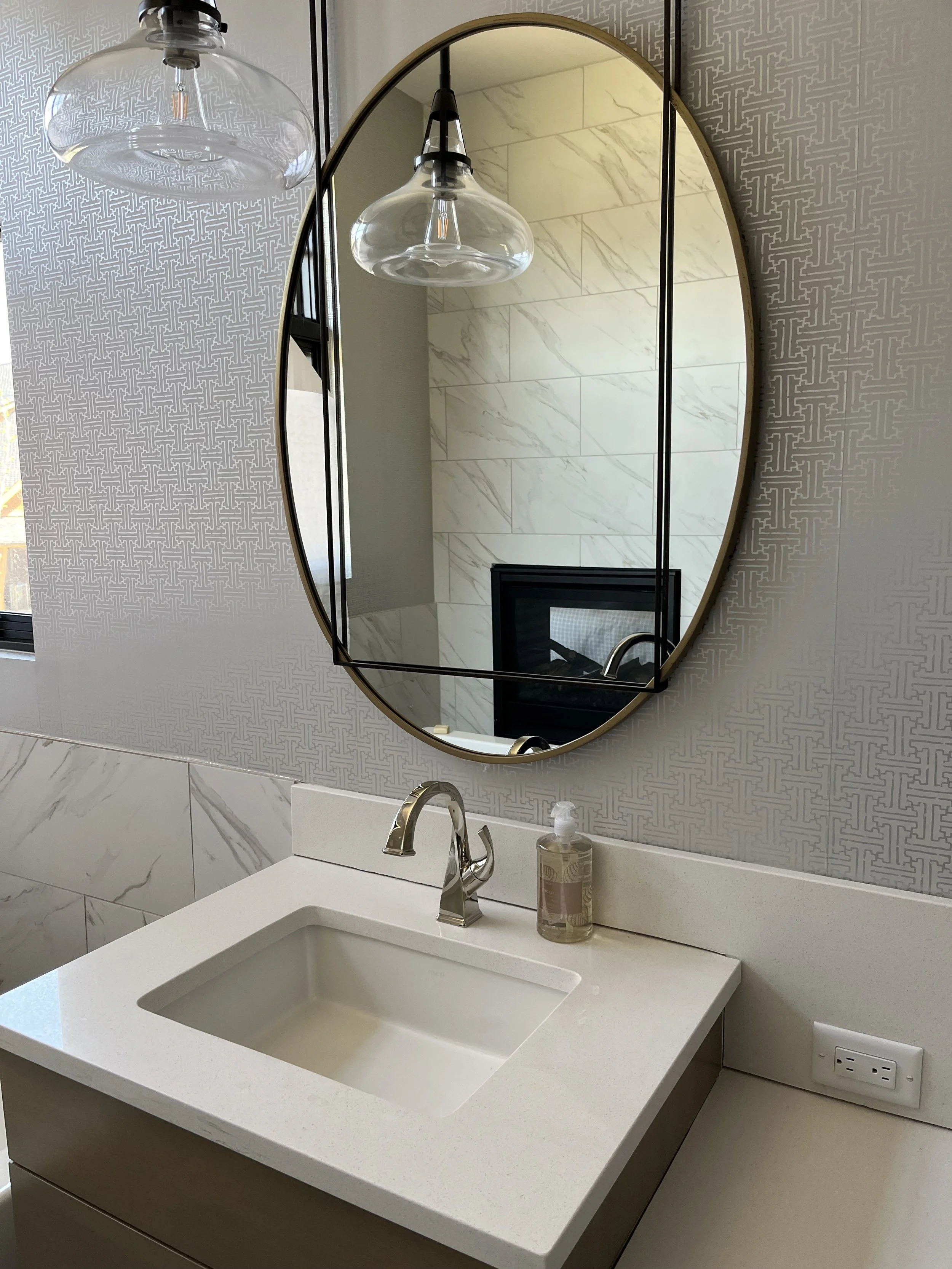 Bathroom with a white countertop sink, a gold faucet, a soap dispenser, and a large oval mirror. There is a pendant light above the mirror, a window on the left, and a patterned wallpaper behind the mirror.