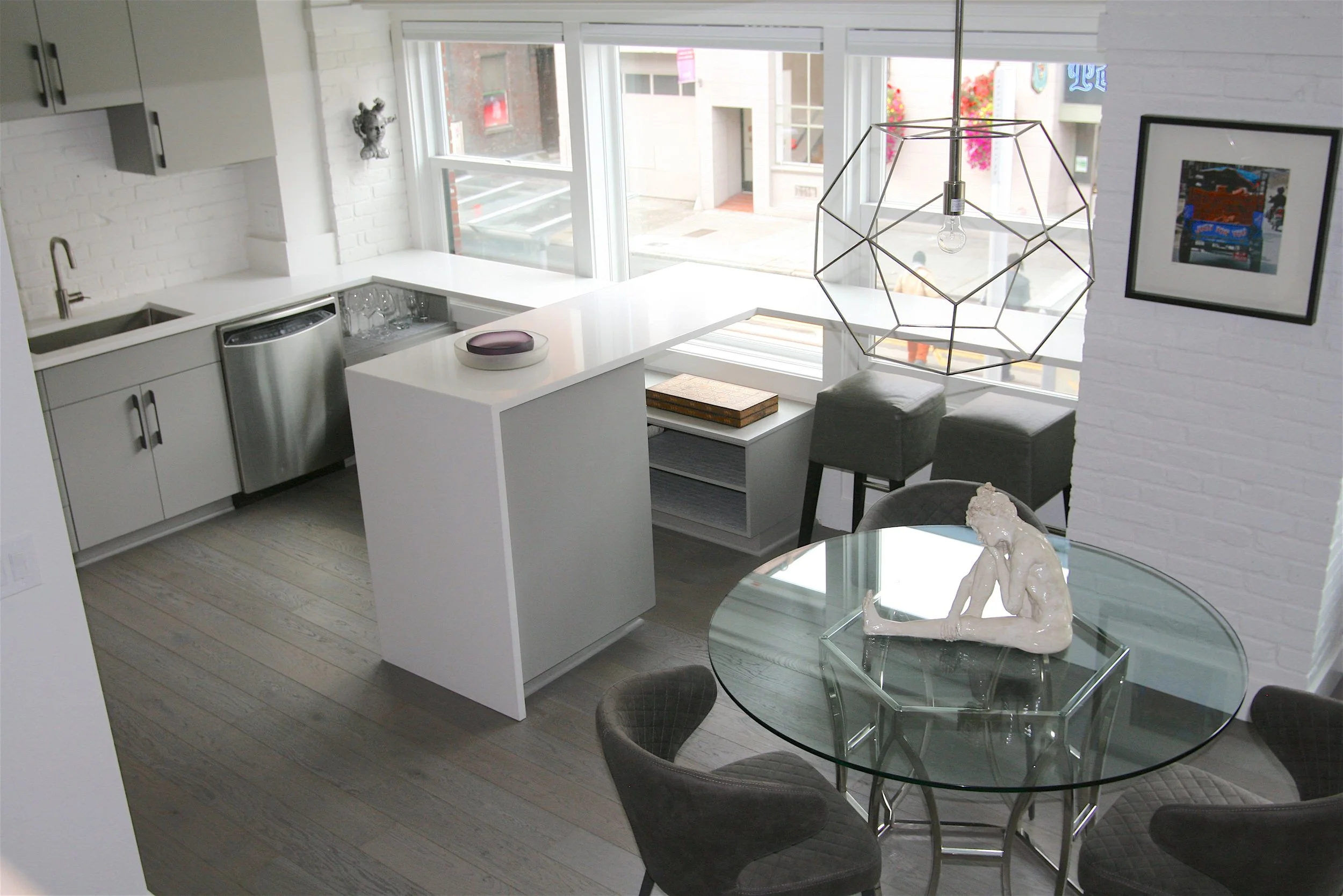 Modern kitchen with white cabinets, a white brick backsplash, and large windows. There is a round glass dining table with four chairs, a white sculpture of a kneeling woman, and a geometric metal pendant light hanging above the table.