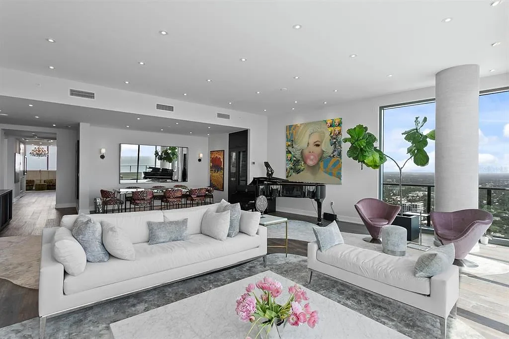 Modern living room with white sofas, colorful artwork, large windows showing city view, and a black grand piano.