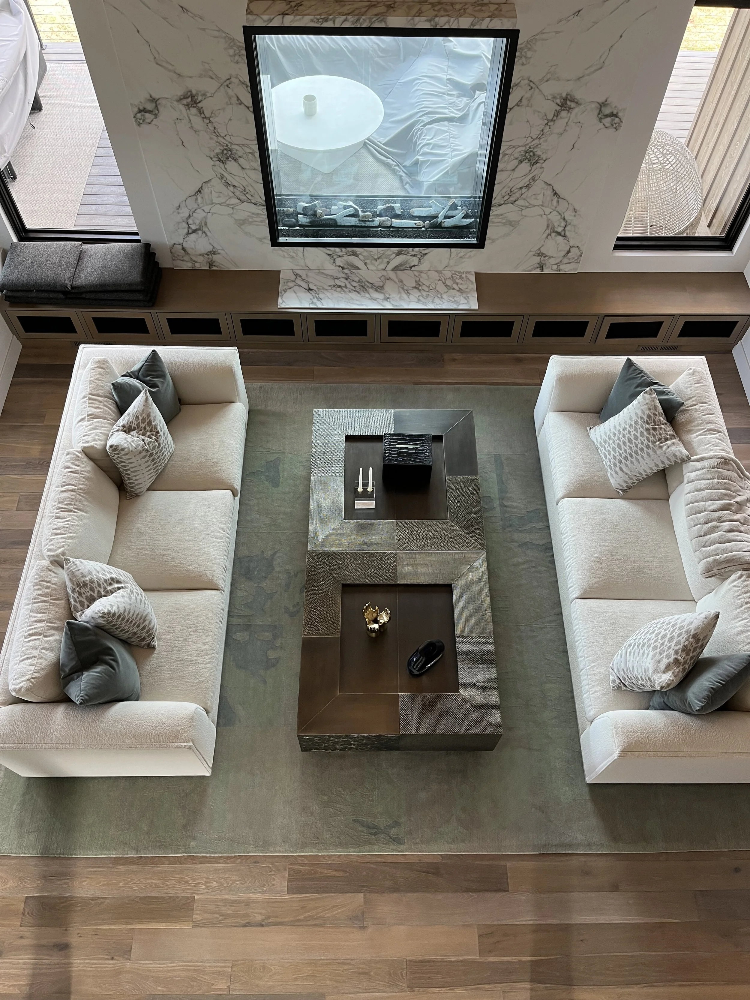 Living room with two white sofas with throw pillows, a central coffee table with decorative items, a large area rug, built-in cabinets beneath a marble wall, a window with patio doors showing an outdoor deck, and a marble fireplace. The photo is take