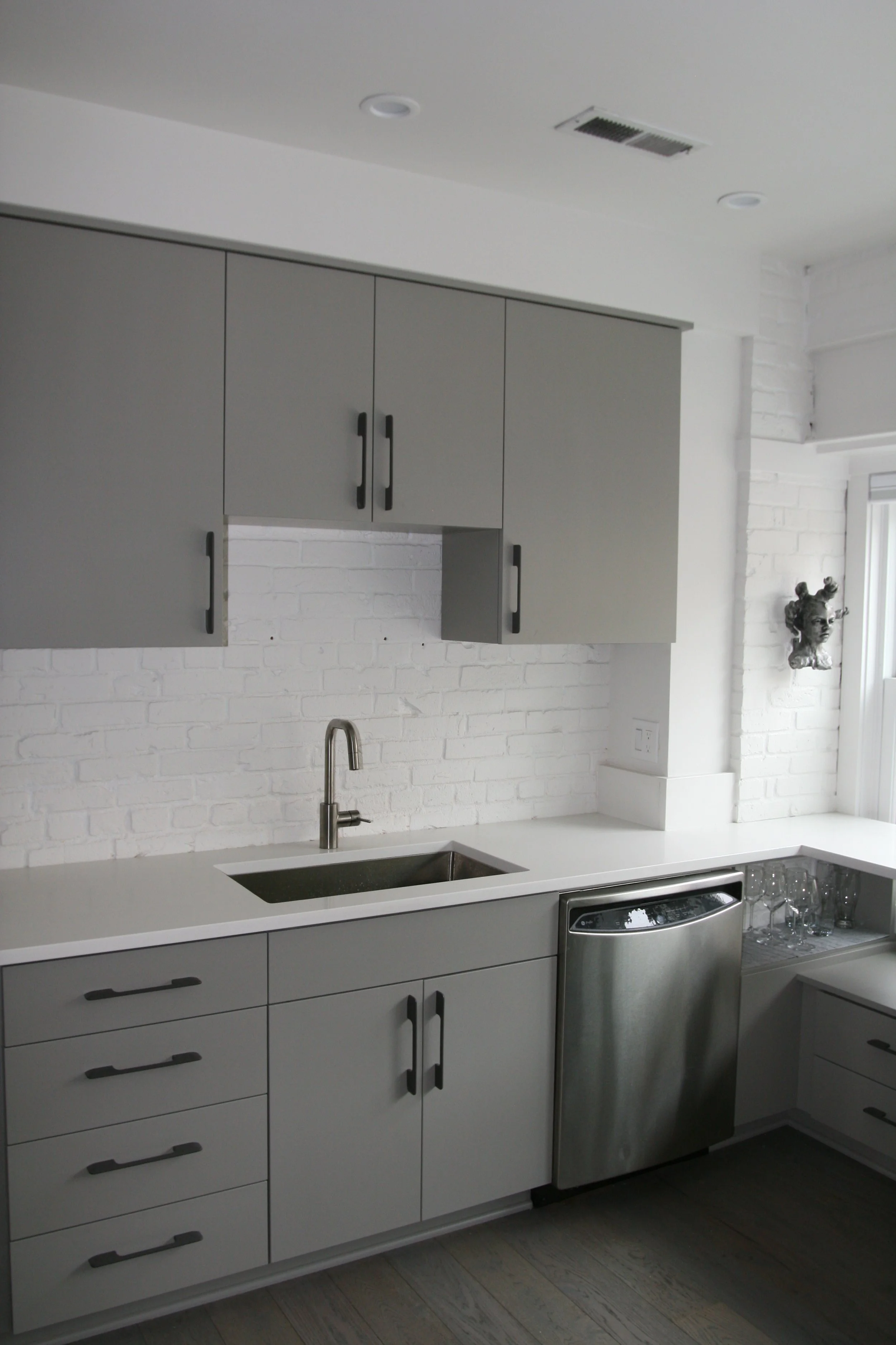 Modern kitchen with grey upper cabinets, white lower cabinets, a stainless steel dishwasher, a sink with a single-handle faucet, a white brick backsplash, and decorative wall art sculpture of a woman's head.