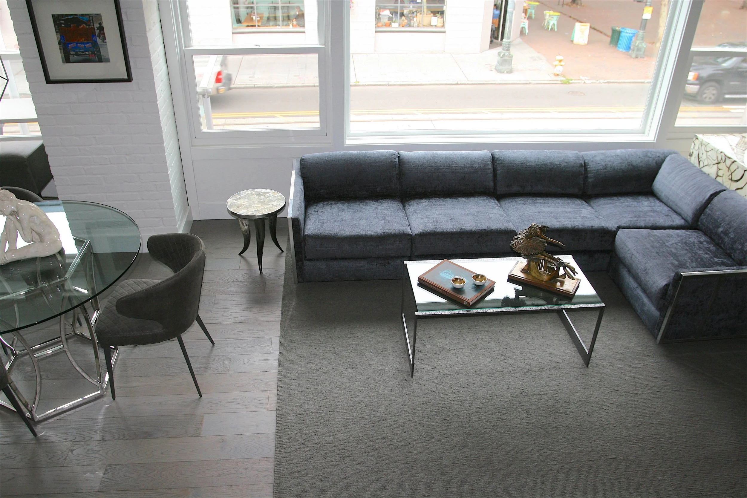 Interior view of a modern living room with large windows, a dark gray sectional sofa, glass-top coffee table with decorative items, a small side table, and dining area with a glass table and black chairs. Outside the windows, a street with parked car