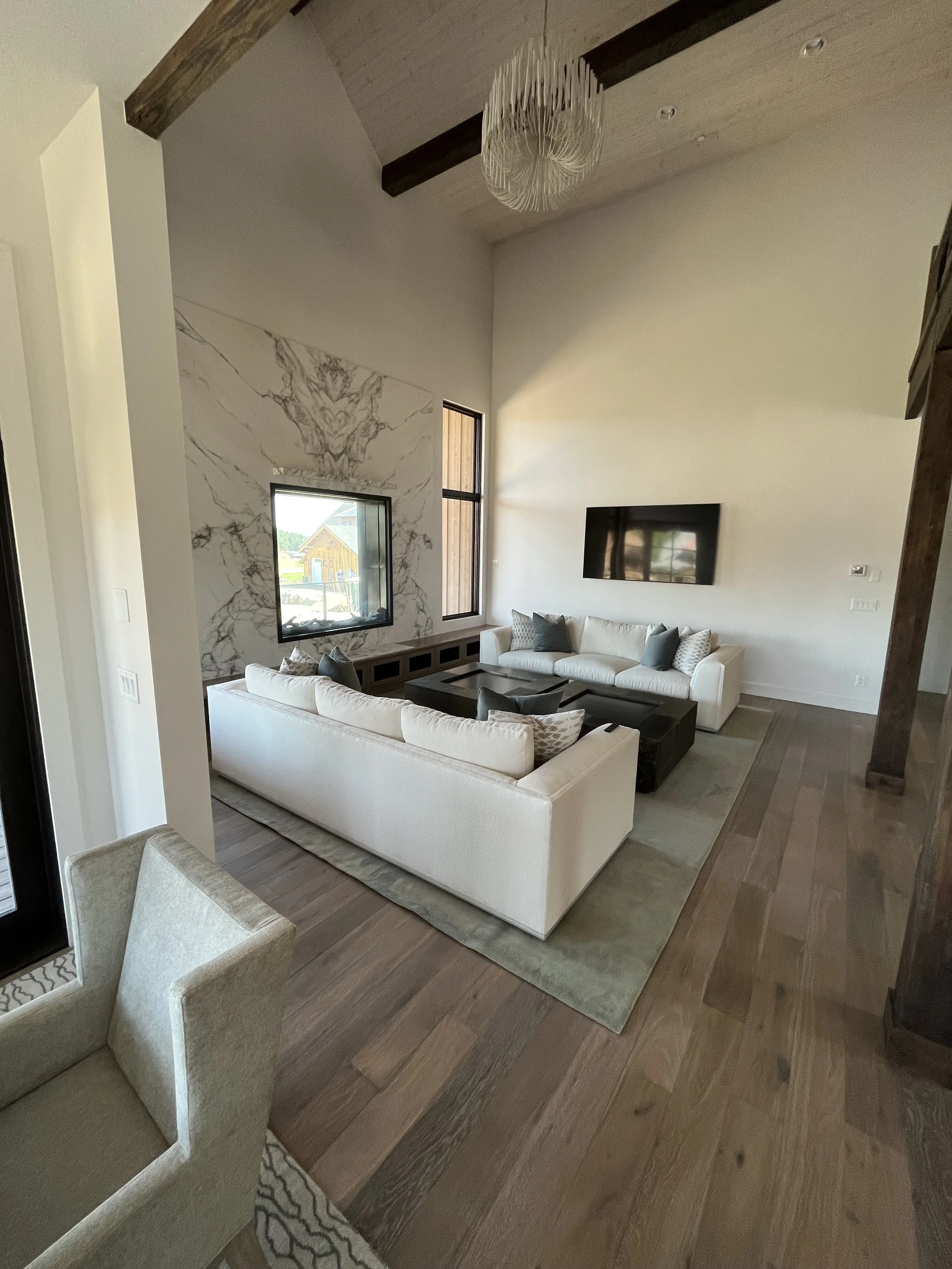 Modern living room with two white sofas, a black coffee table, a large TV on the wall, and a marble accent wall with windows and a wooden beam ceiling.