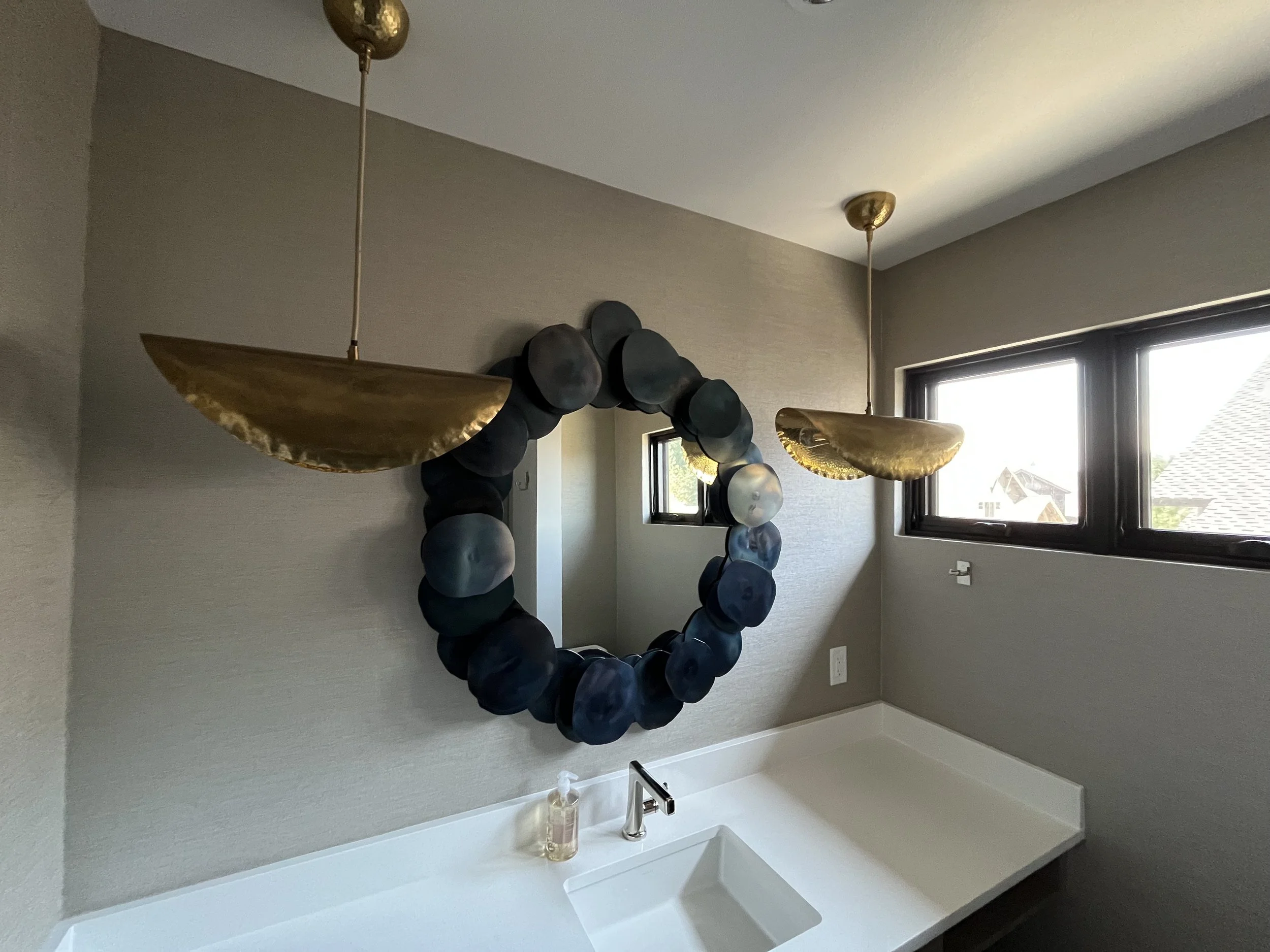 Bathroom with a white countertop sink, a framed mirror decorated with dark circular objects, grey walls, and two large windows letting in natural light.