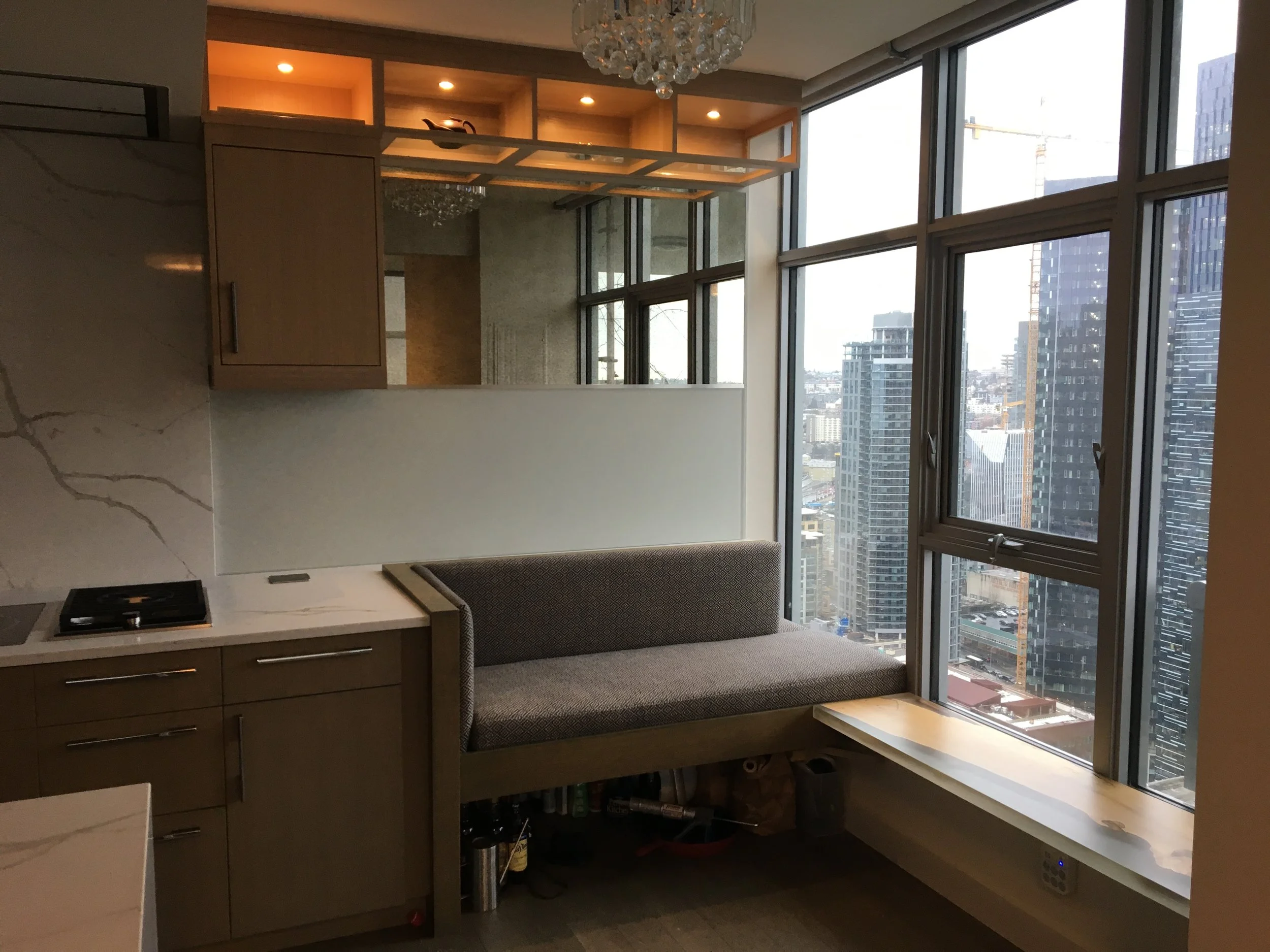View from inside a high-rise apartment with large windows showing a city skyline of tall buildings, built-in kitchen cabinets, and a small sitting area with a bench.