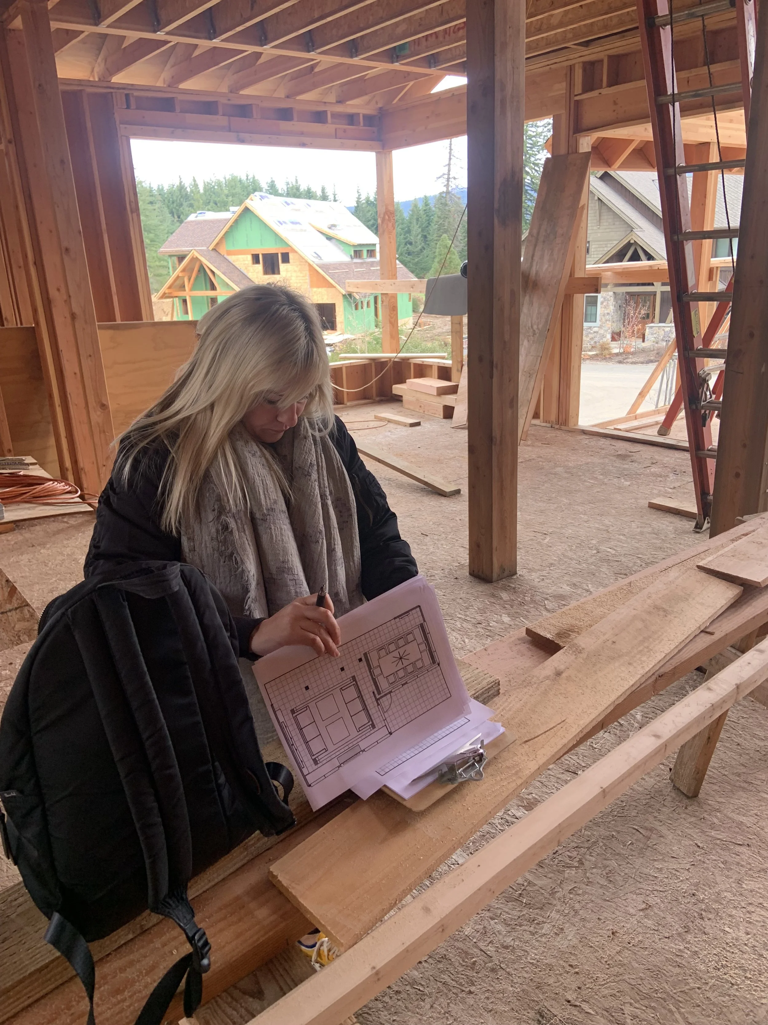 A woman with blonde hair wearing a black jacket and scarf is holding architectural plans while standing in a wooden house under construction. She is in front of a workbench with wooden planks and a backpack with a camera strap is visible.