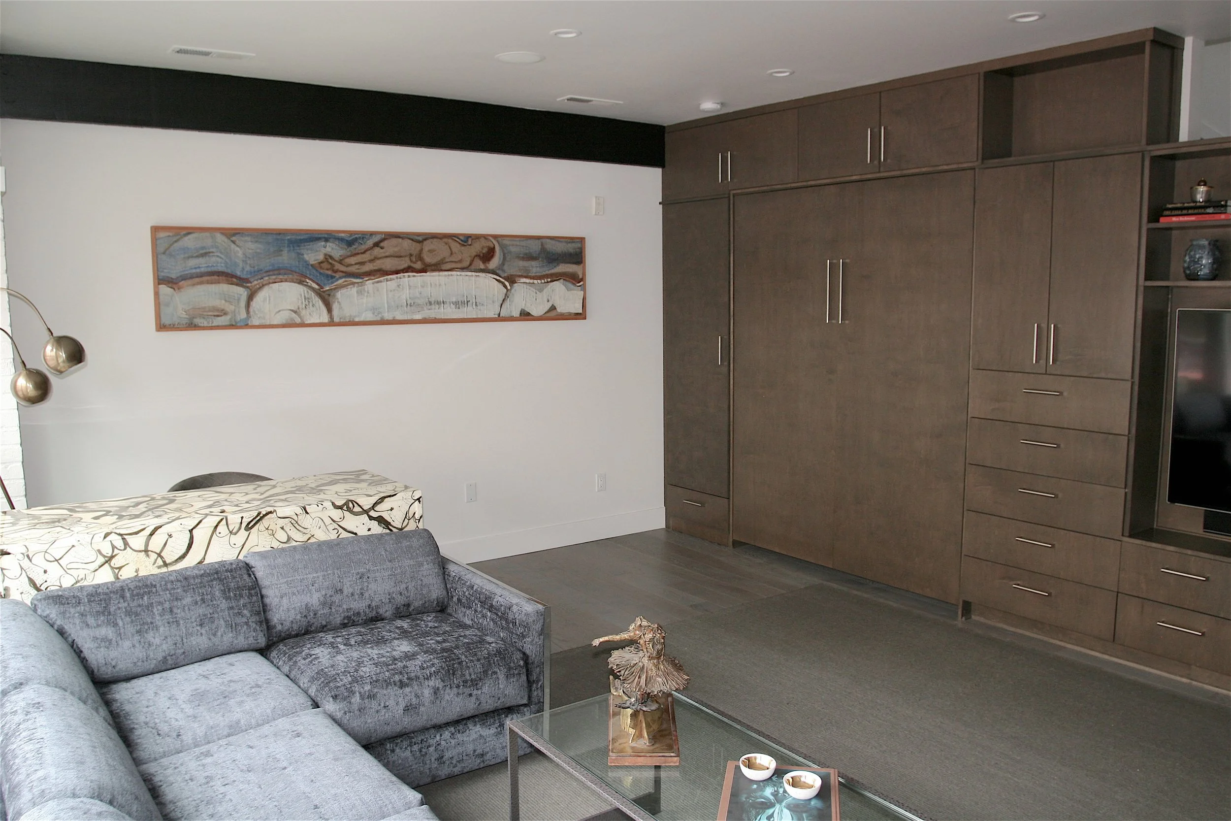 Living room with a grey sofa, a glass coffee table with decorative items, a beige area rug, a modern wooden wall unit, an abstract painting on the white wall, and pendant lighting fixtures.
