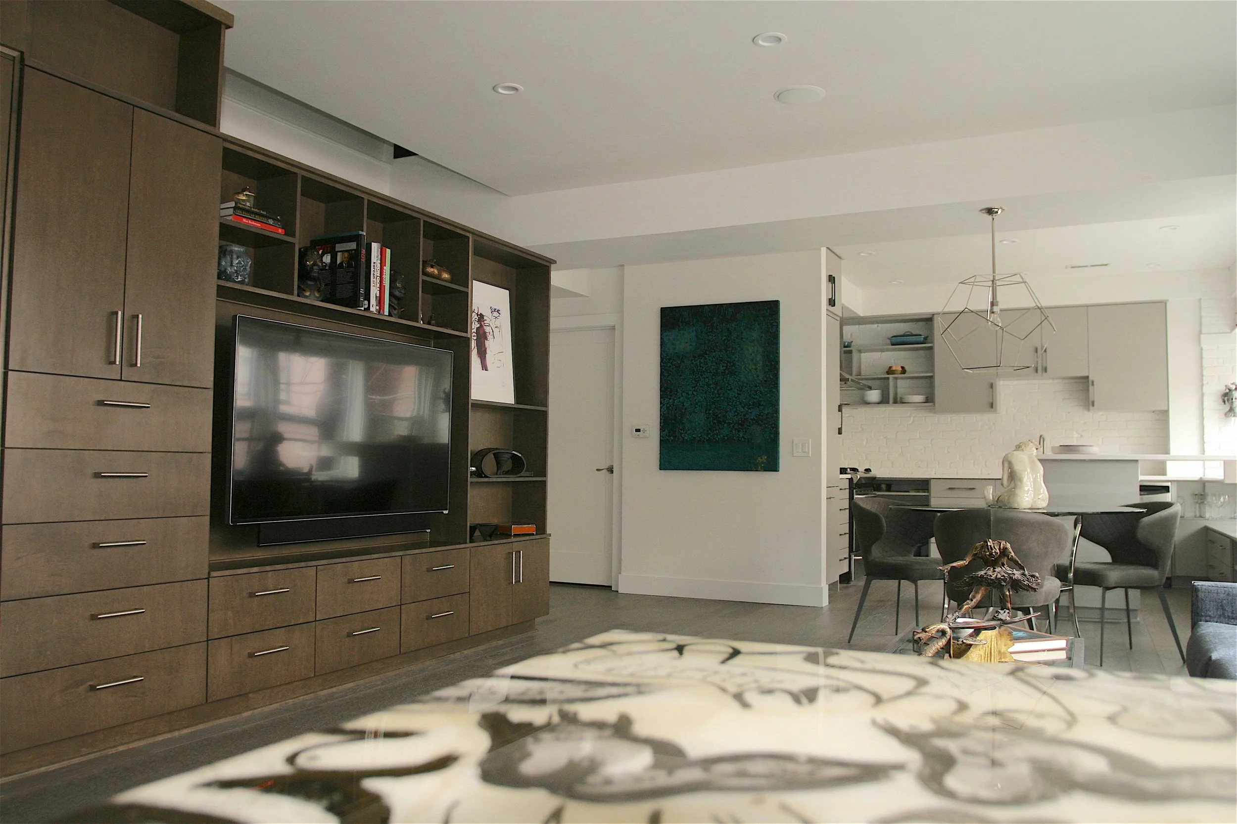 Modern open-plan living room and kitchen with wooden cabinetry, a large flat-screen TV, and a sitting area with chairs and a decorative sculpture on the table.