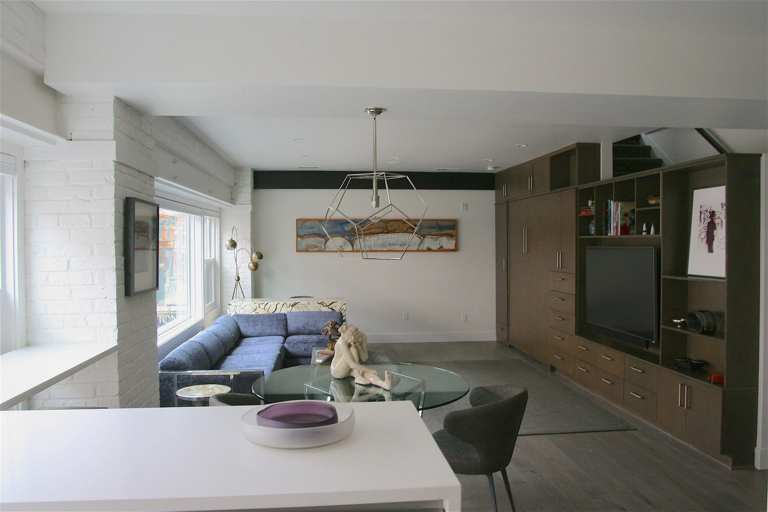 A modern living room with white brick walls, large windows, and a gray sofa. A dining table with black chairs and decorative items such as a sculpture and purple bowl are visible. There is a ceiling light fixture, a framed painting, and built-in dark