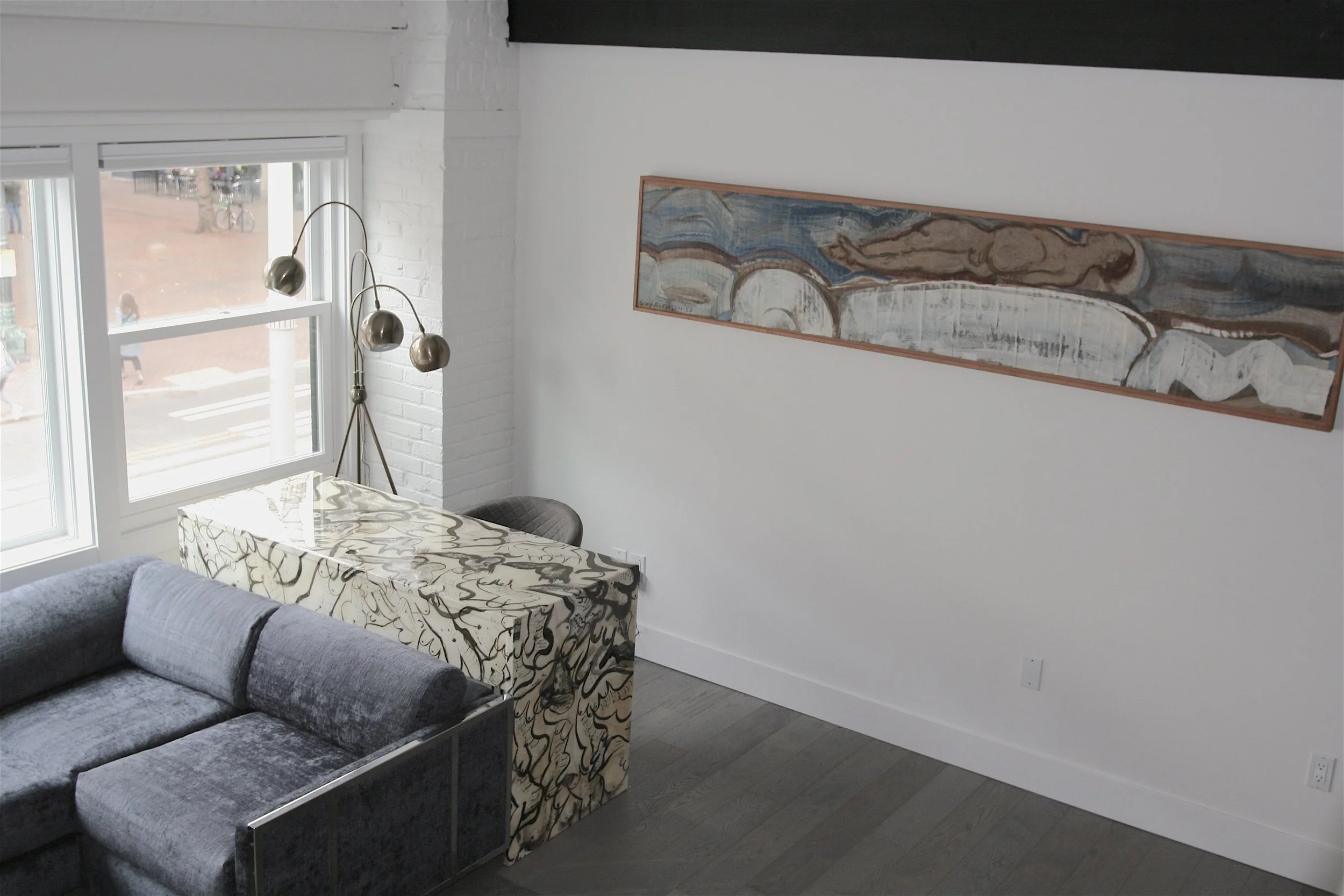A corner of a modern living room with a gray sofa, a patterned armchair, a white brick wall, a large window, a tall floor lamp, and an abstract painting hanging on the wall.