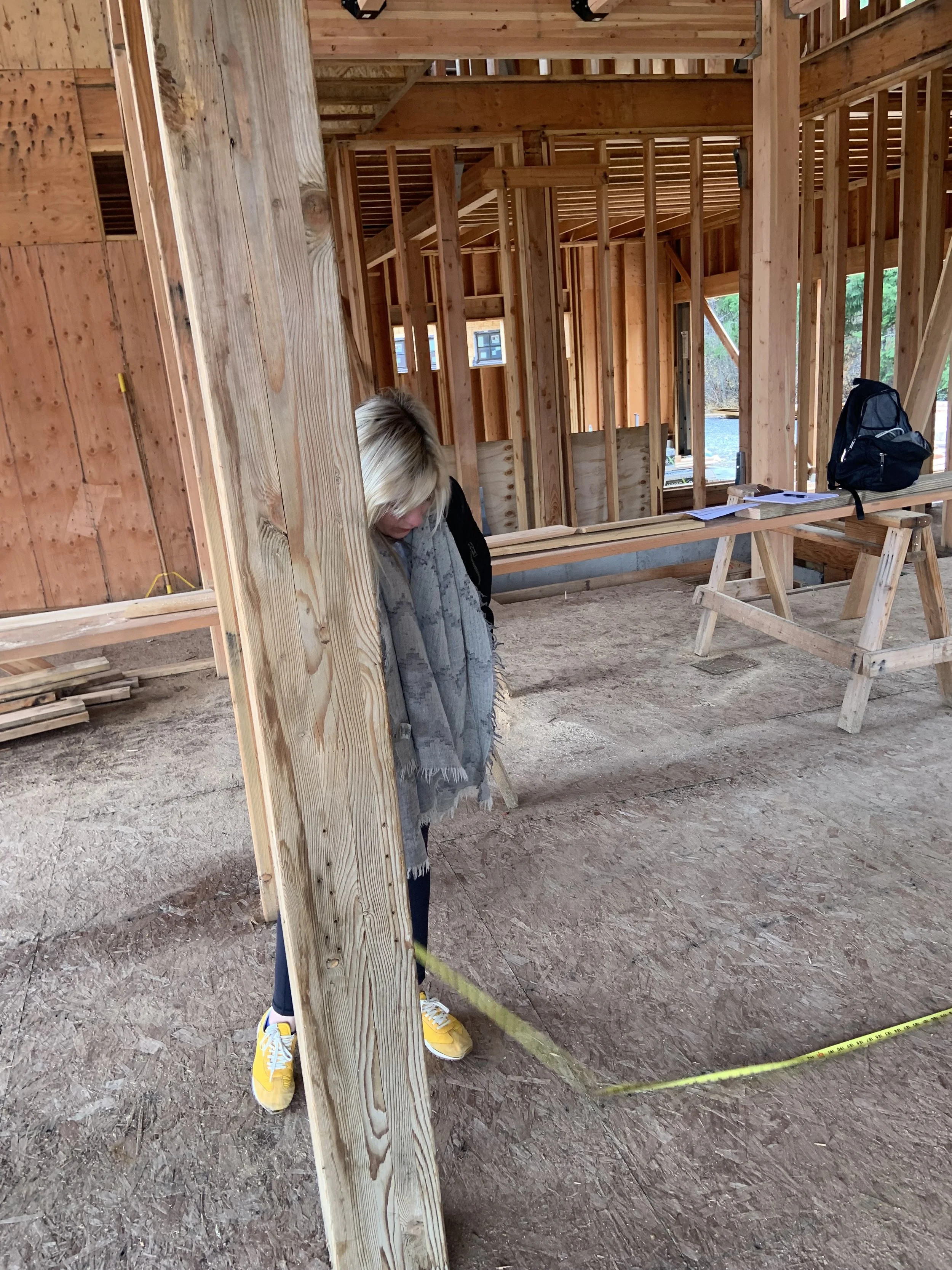 A woman with blonde hair wearing yellow sneakers and dark pants stands inside a wooden frame construction site. She is holding a measuring tape and looking down. The construction site has exposed wooden studs and beams, with some planks and construct