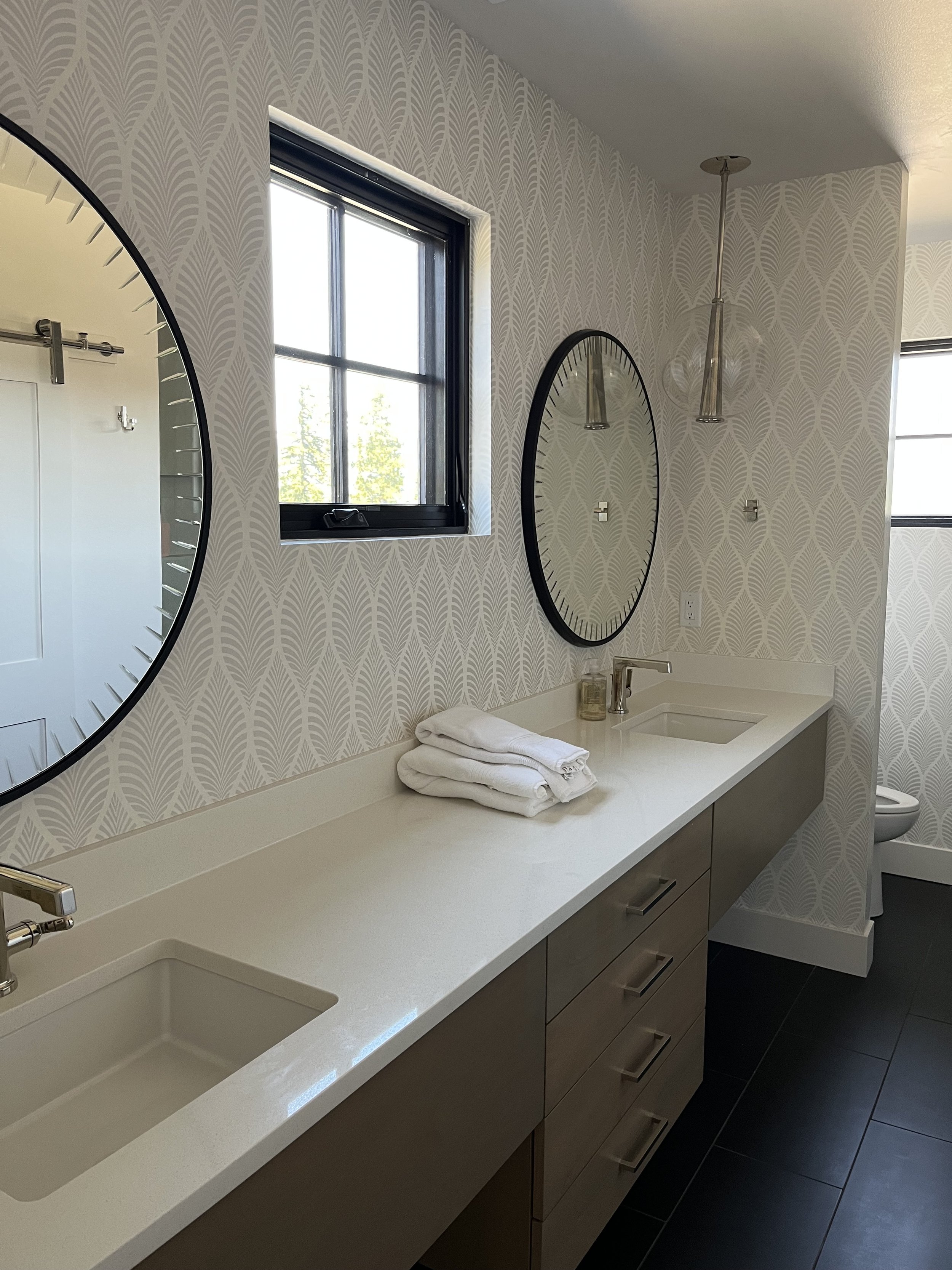 Modern bathroom with dual sinks, large mirror, patterned wallpaper, black-framed window, hanging pendant lights, and folded towels on the countertop.
