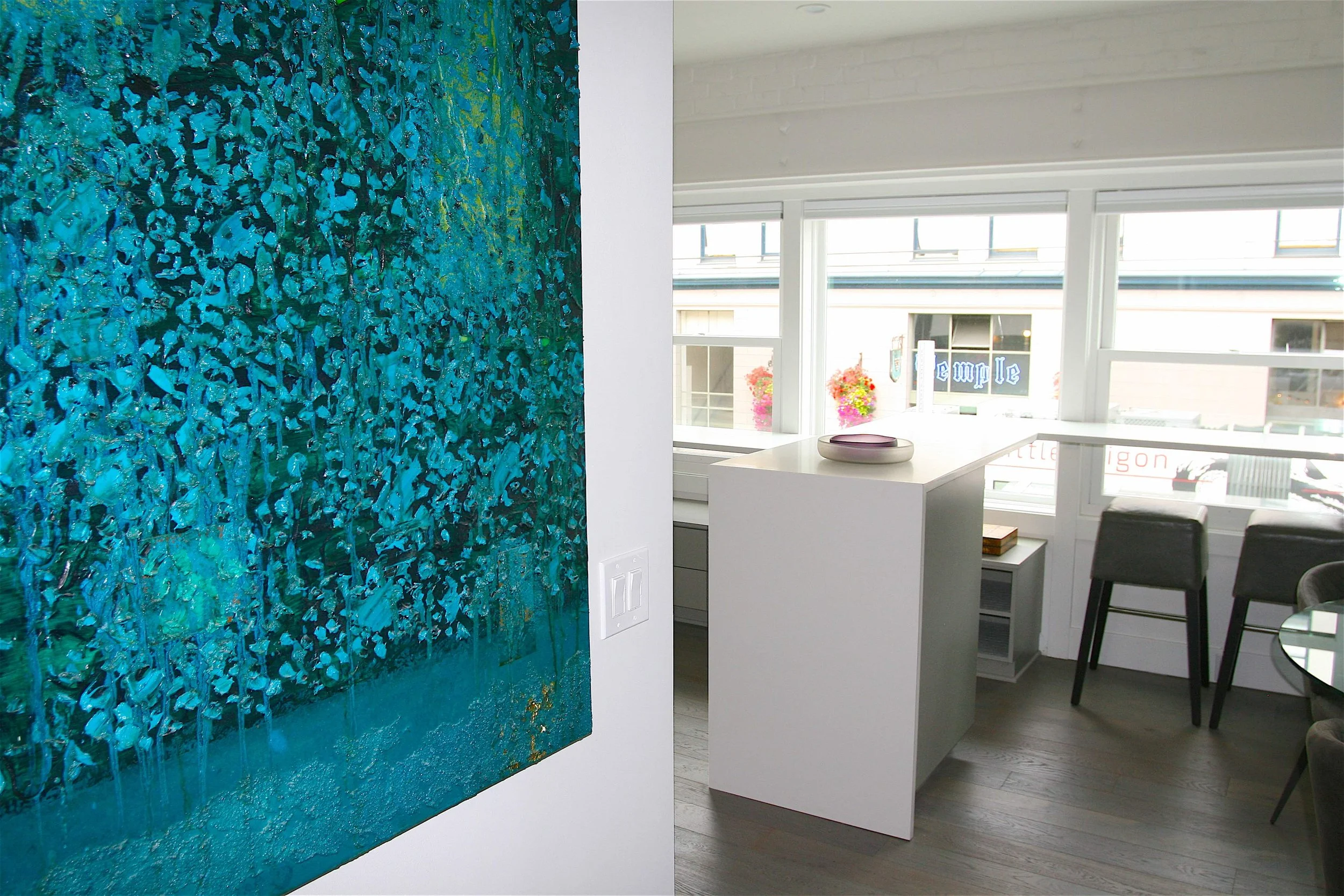 A split view of an art gallery: on the left, a large textured painting with shades of blue, teal, and some gold accents; on the right, a bright city view through large windows with a white interior, white countertop, and black chairs.