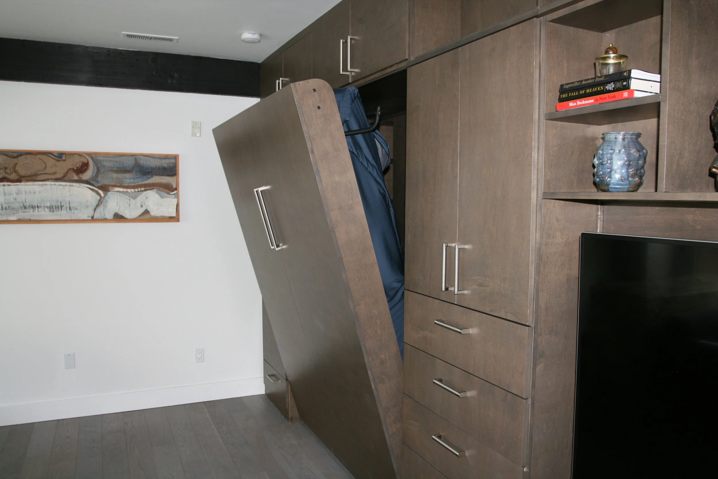 View of a modern kitchen or office space with dark brown wooden cabinets and a matching wooden desk. A grayish-blue backpack rests on the desk. Adjacent to the desk is built-in shelving containing books, a decorative jar, and a small container. An ab