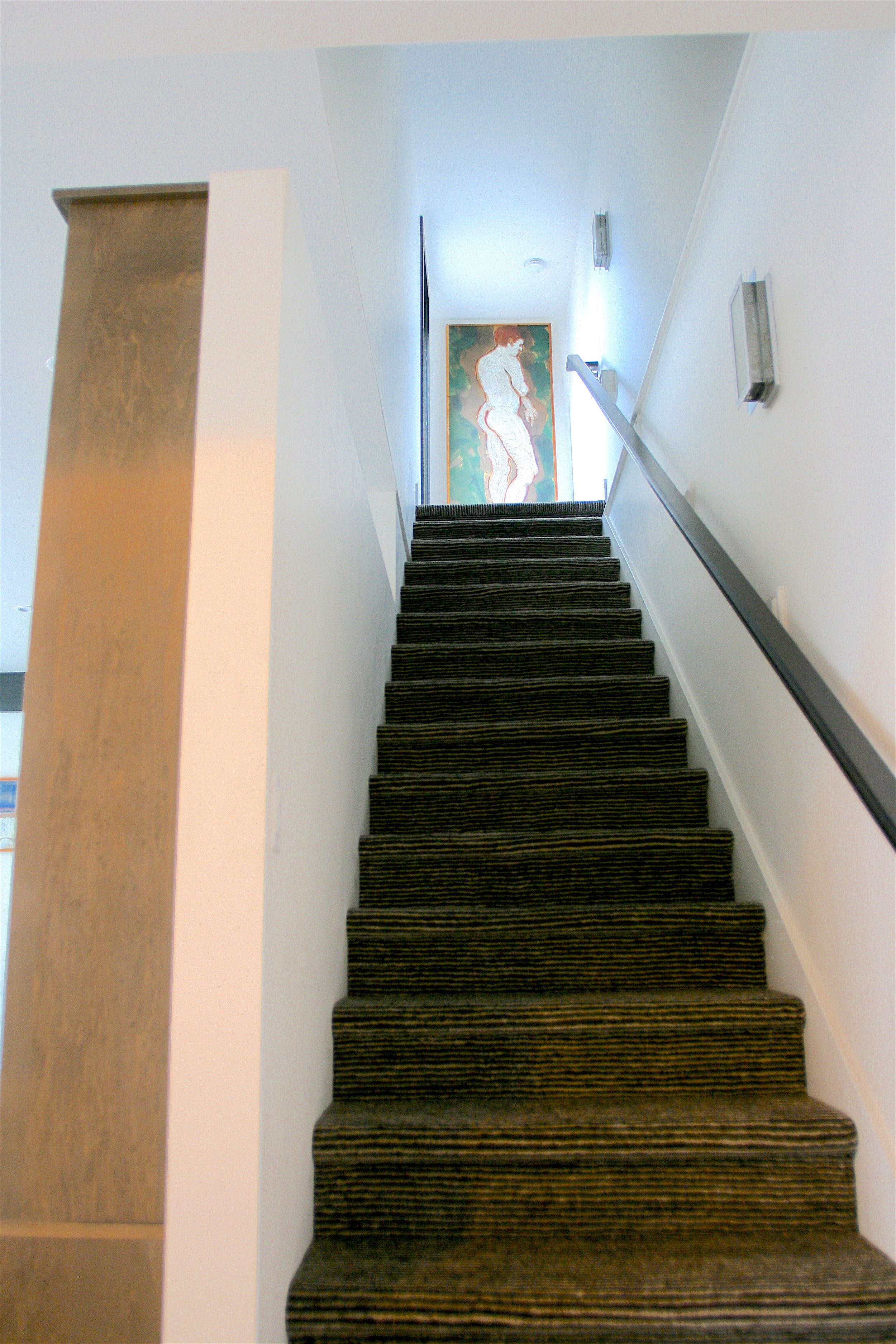 Interior view of a staircase with striped carpet, white walls, and a handrail on the right. A colorful framed artwork of a woman is at the top of the stairs near a bright window or light fixture.