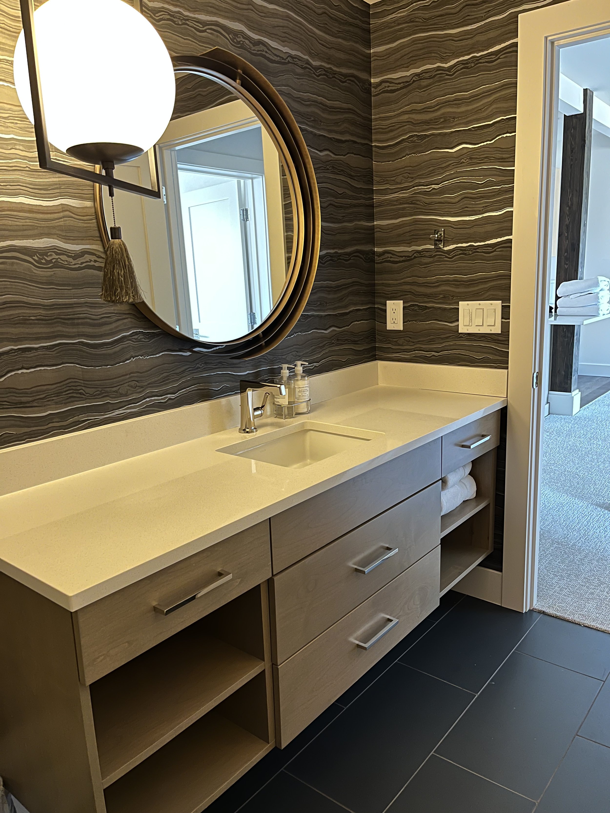 Bathroom vanity with a cream countertop, a built-in sink, a modern faucet, a mirror, wall-mounted light fixture, and towels stored in open shelves below. The wall has a dark, wavy, striped wallpaper, and there are multiple electrical outlets and swit