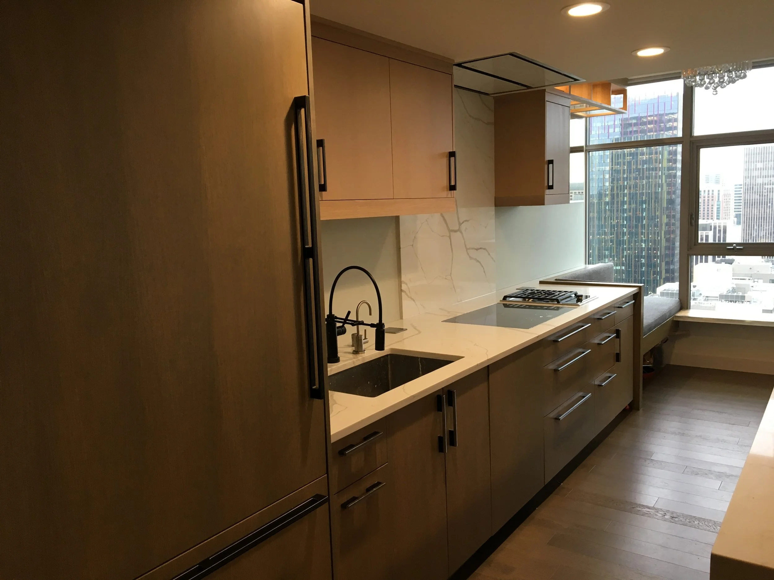 Modern kitchen with wooden cabinets, marble backsplash, and large windows showing a cityscape.