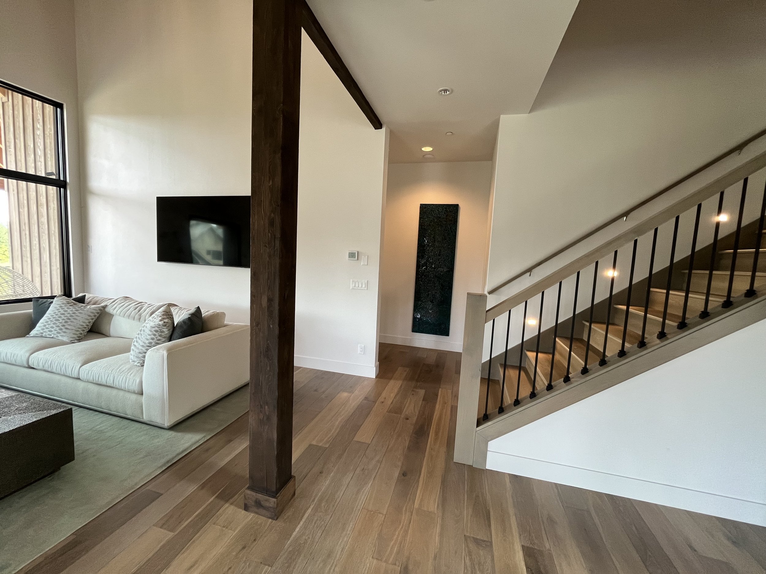 Living room with large window on left, beige sofa with pillows, flat-screen TV on wall, wooden flooring, staircase with black metal railing on right, and a dark vertical artwork on the back wall.