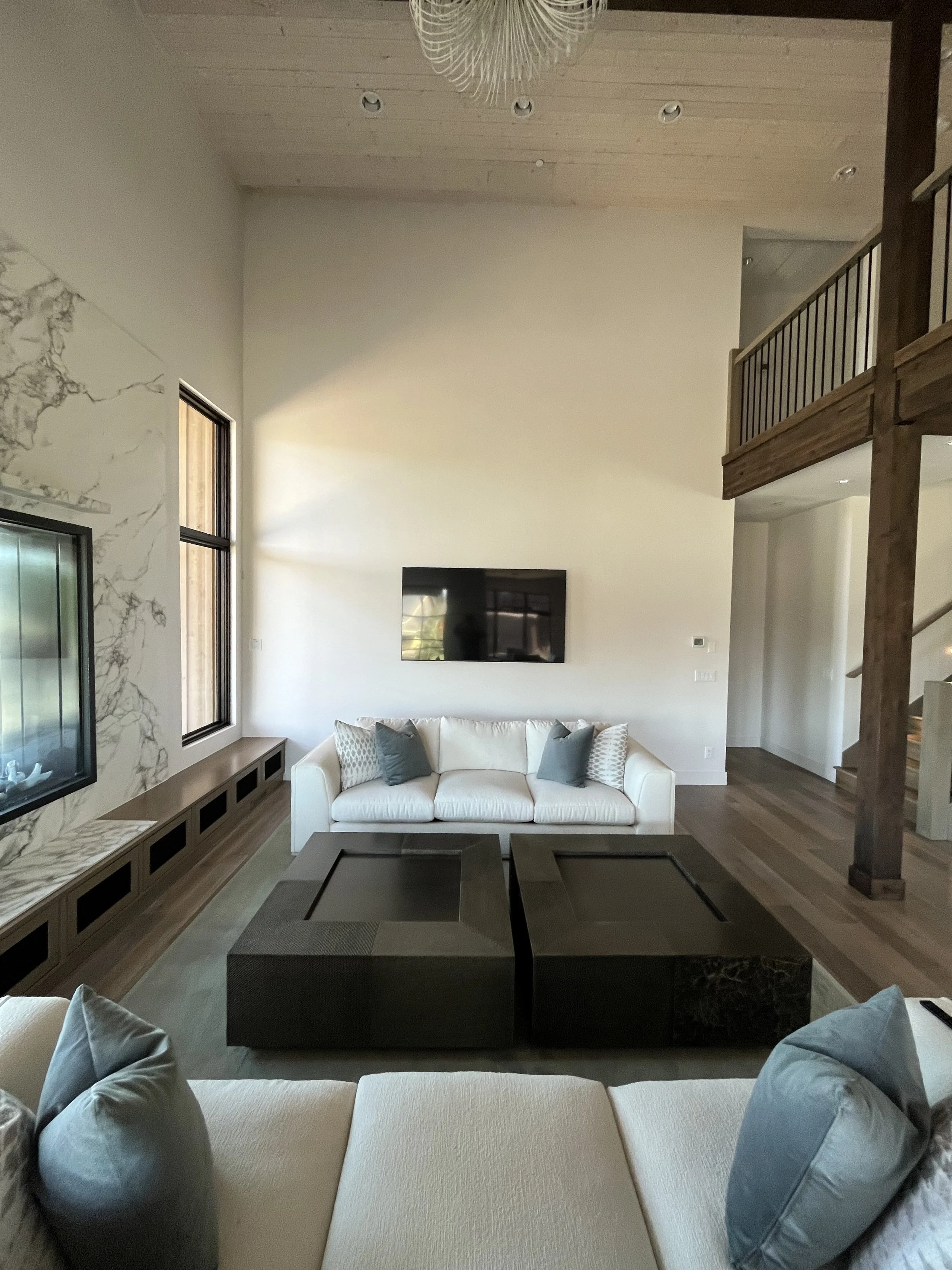 Interior view of a modern living room with a white sofa, gray pillows, a dark coffee table, large windows, and a mounted television on a white wall, with wooden ceiling and stairs.