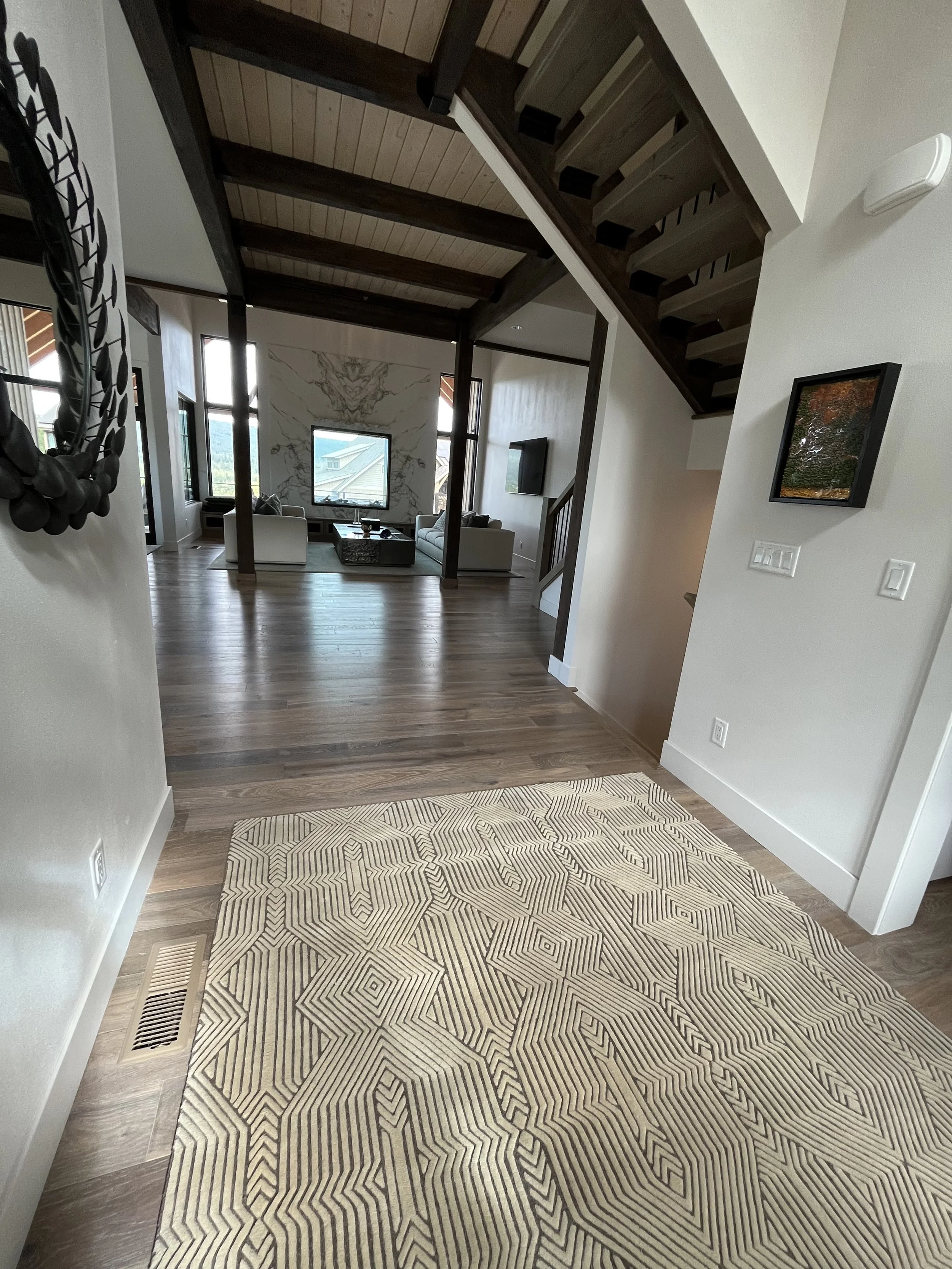 View of a modern living room with wooden flooring, a patterned beige and black area rug, a staircase with dark wood steps, and large windows showing outdoor scenery with mountains and rooftops.