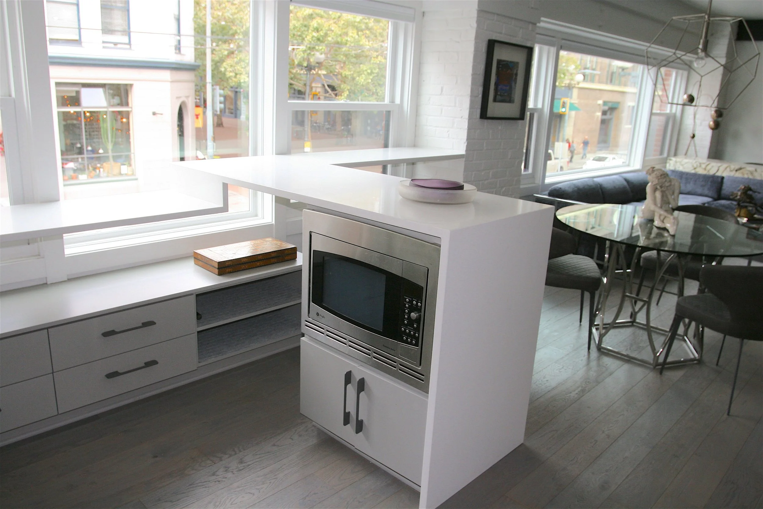 Bright modern apartment interior with large windows, white brick wall, dining area with glass table and chairs, kitchen with microwave, and city street view outside.