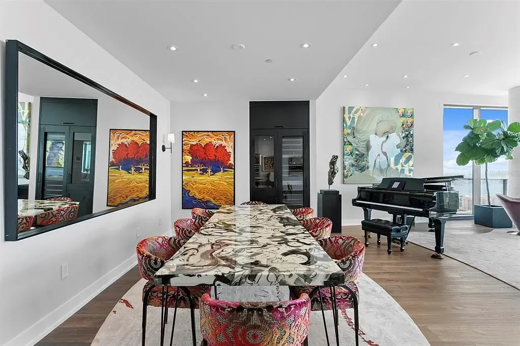 Modern dining room with a long marble table, colorful patterned chairs, and vibrant artwork on the walls. Also features a black grand piano near a large window with a potted plant.
