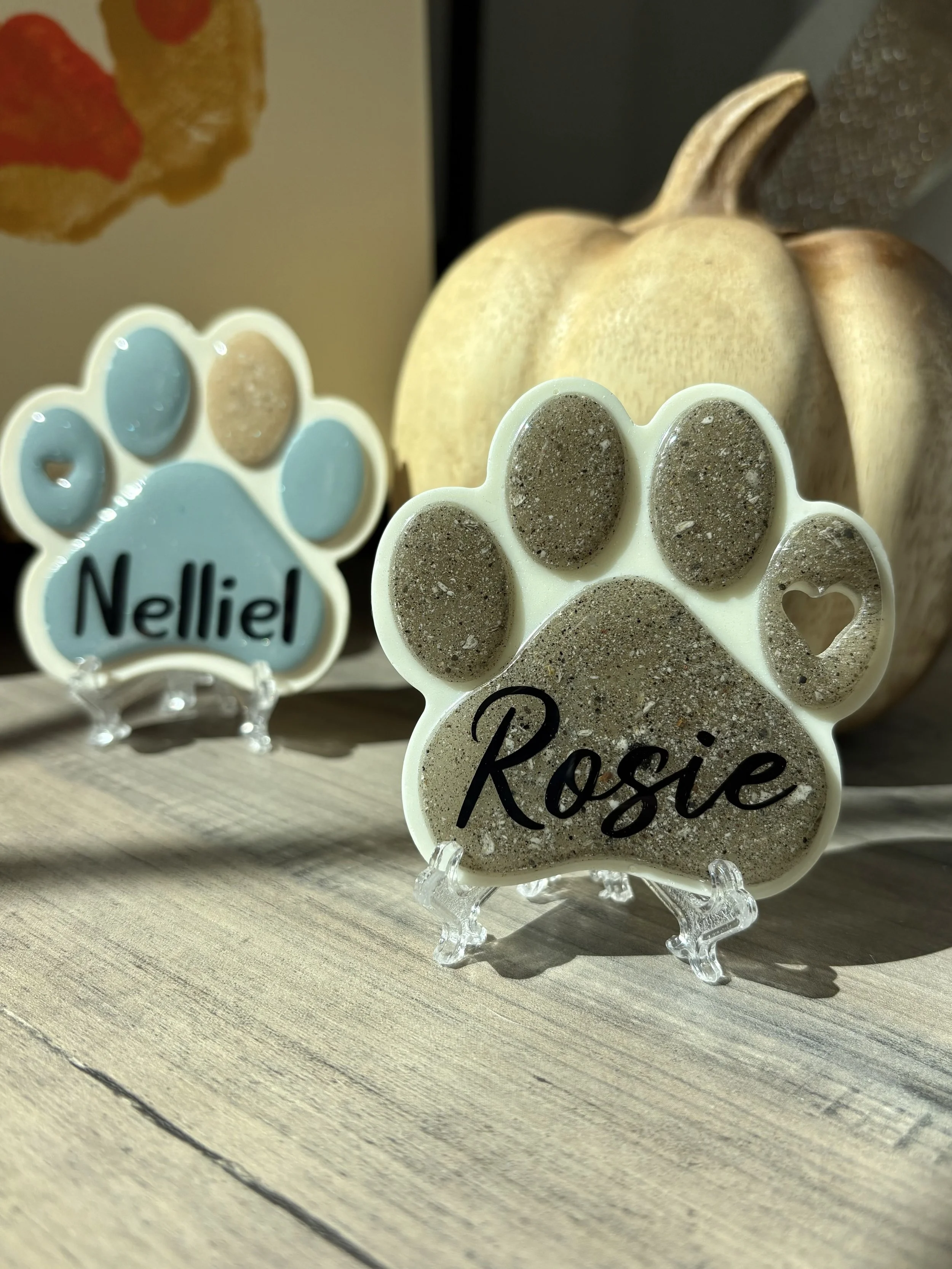 Two paw print-shaped resin keepsakes with pet ash in the toe beans, one with the name 'Rosie' and the other with 'Nelliel,' placed on a wooden surface near a wood pumpkin, with a blurred background.