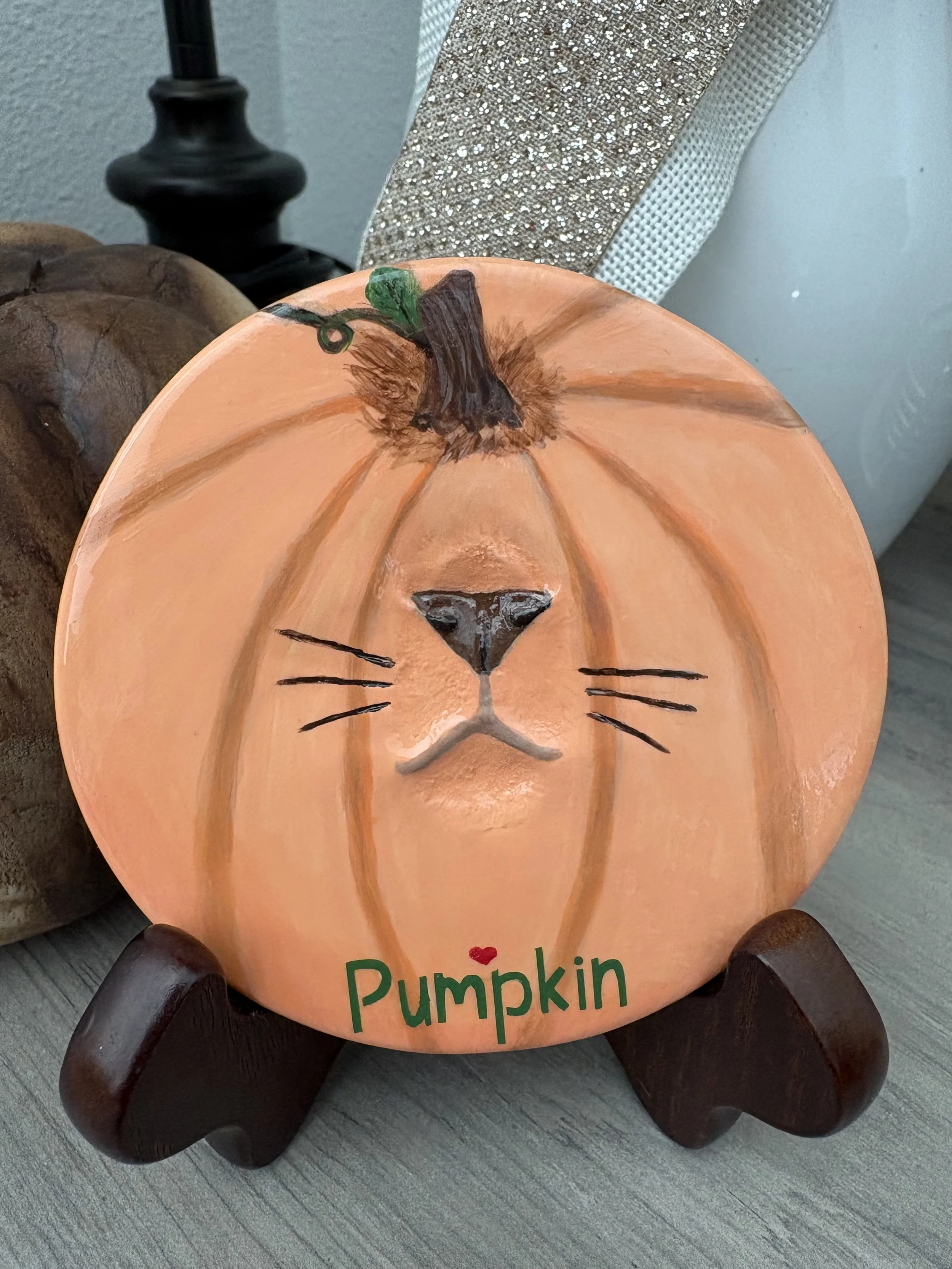 Decorative cat nose impression, painted pumpkin face, resting on a wooden stand with a black lamp and a textured silver surface in the background.