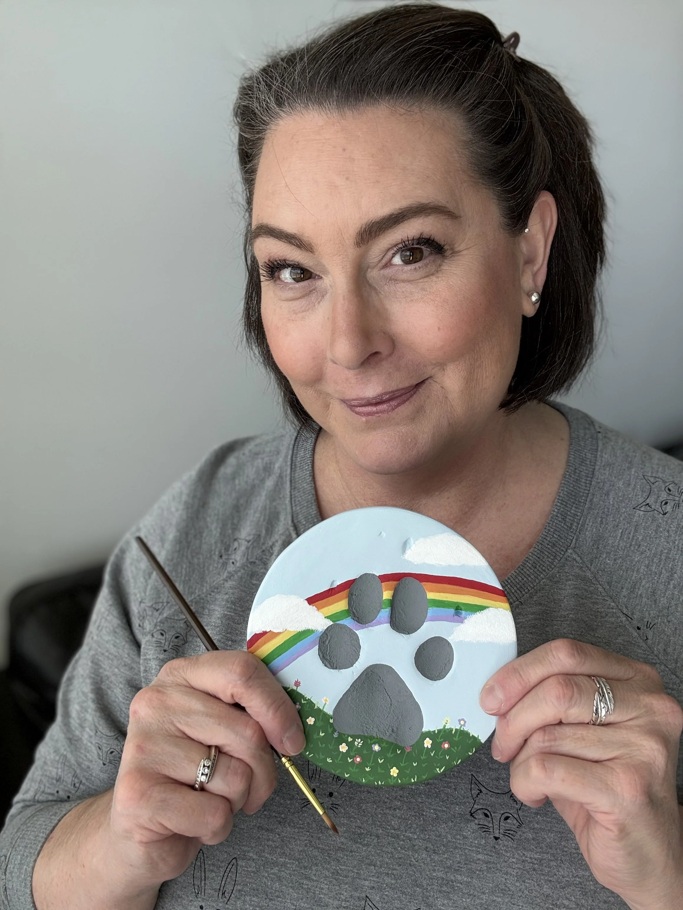 A business owner woman with dark hair and earrings holding a painted paw impression with a rainbow, grass field with flowers, and cloud design.