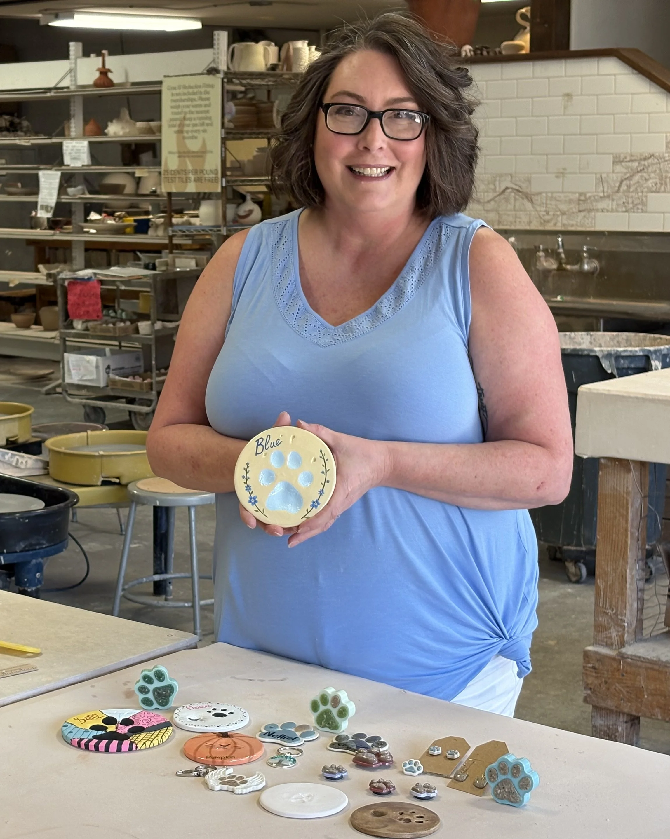 A business owner woman with glasses and curly hair smiling and holding a decorated dog paw impression in a pottery studio. The background features shelves with pottery items and a white brick wall. There are pet memorial products on the table.