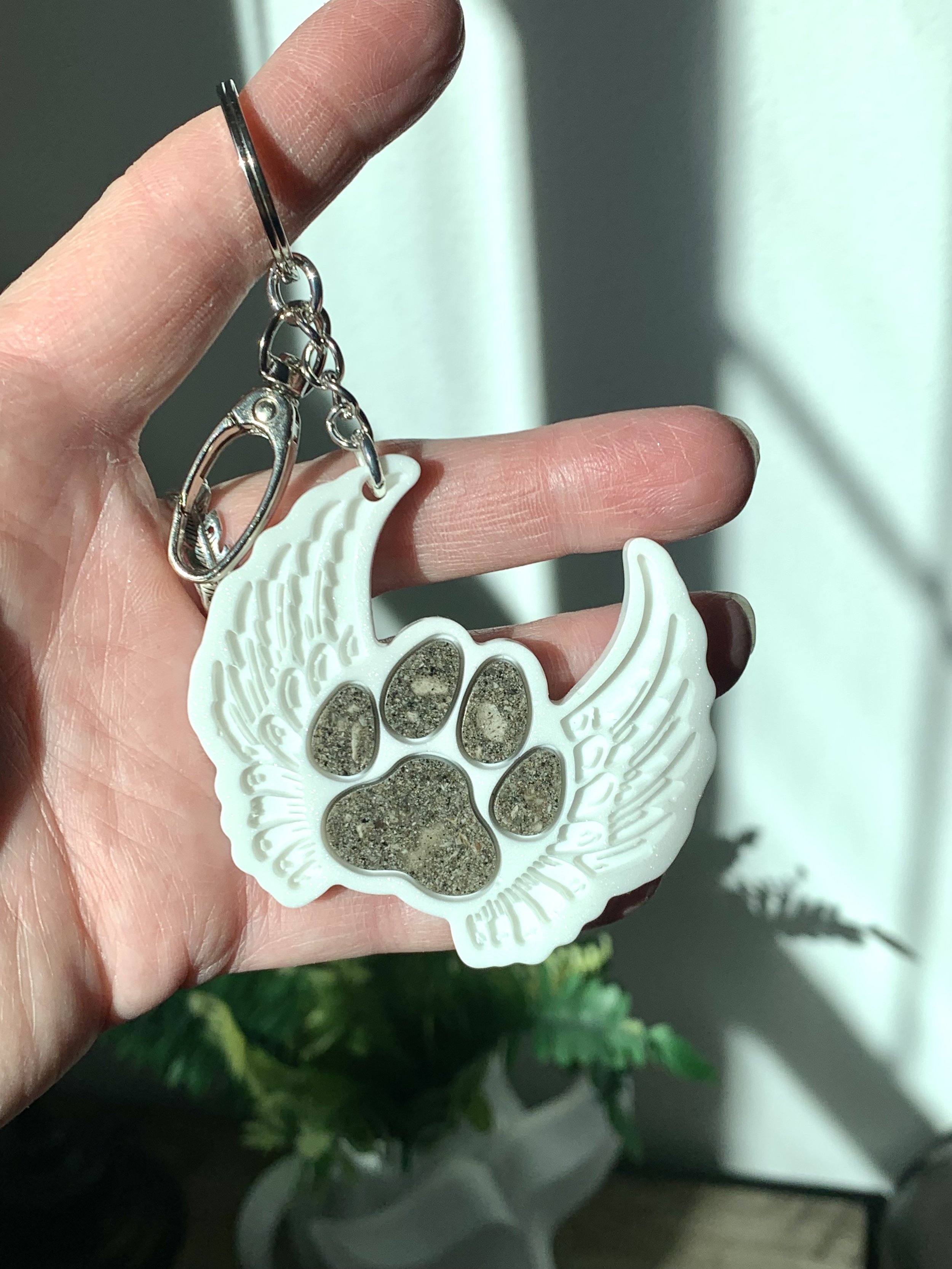 A person holding a white resin keychain with a paw print design filled with pet ash. The background shows some greenery and a blurred setting.